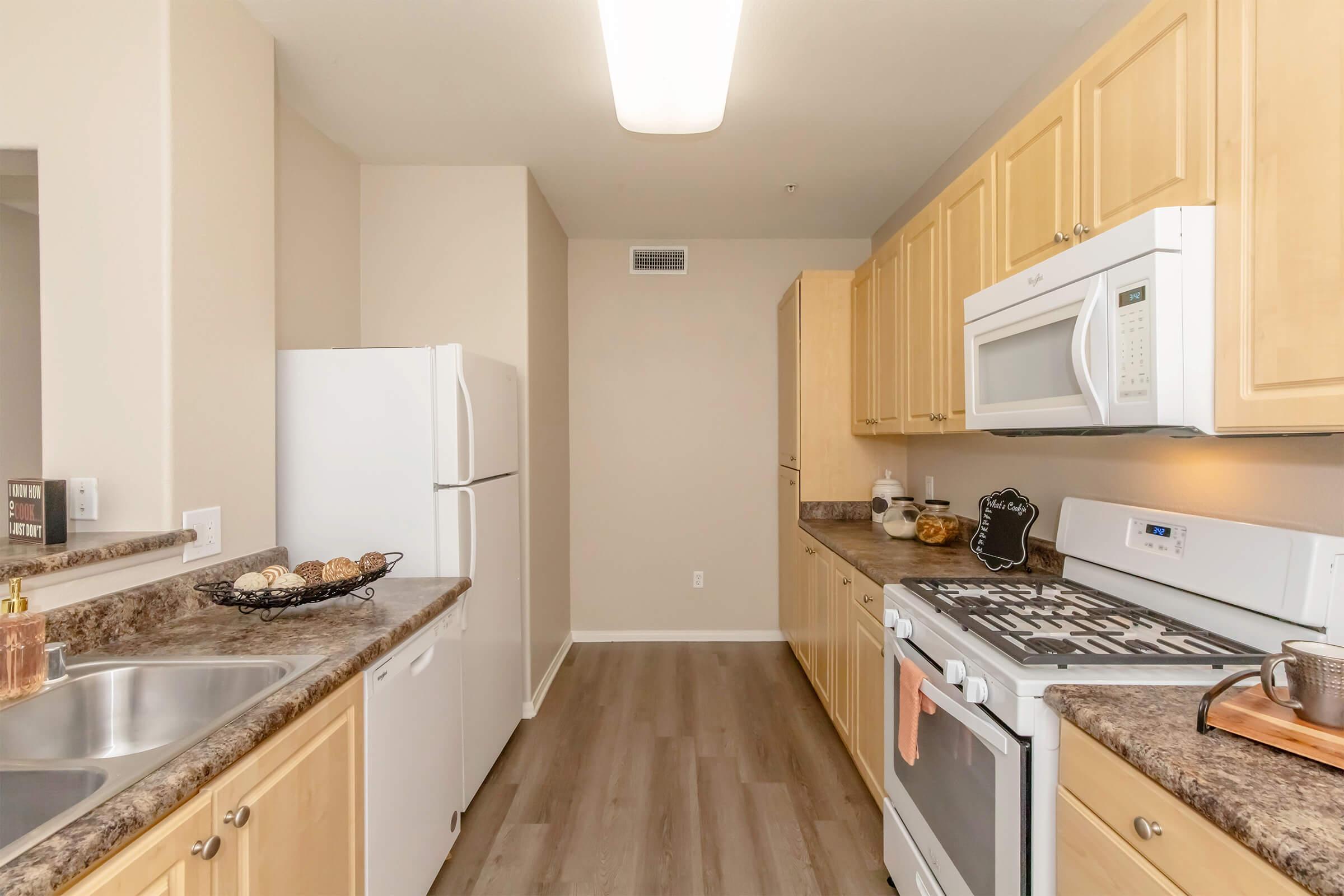Modern kitchen featuring light wood cabinetry, stainless steel appliances, and a spacious countertop. The layout includes a refrigerator, a microwave, and a gas stove. Natural light fills the space, enhancing the neutral color scheme and providing a warm, inviting atmosphere.