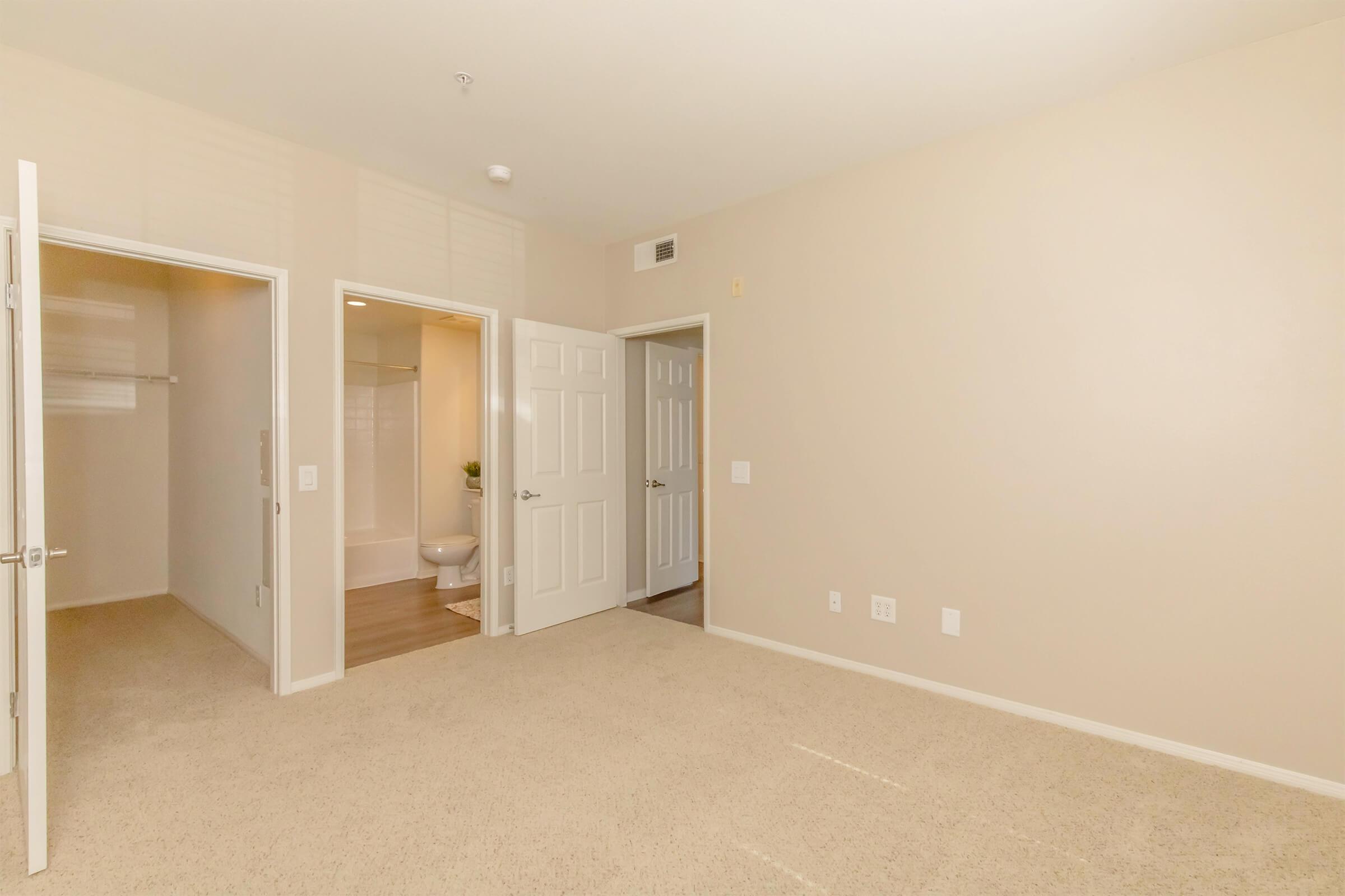 A clean, empty room with beige walls and carpet. Two open doors lead to closets, and a third door leads to a bathroom. The natural light illuminates the space, showcasing a spacious layout with no furniture or decorations.