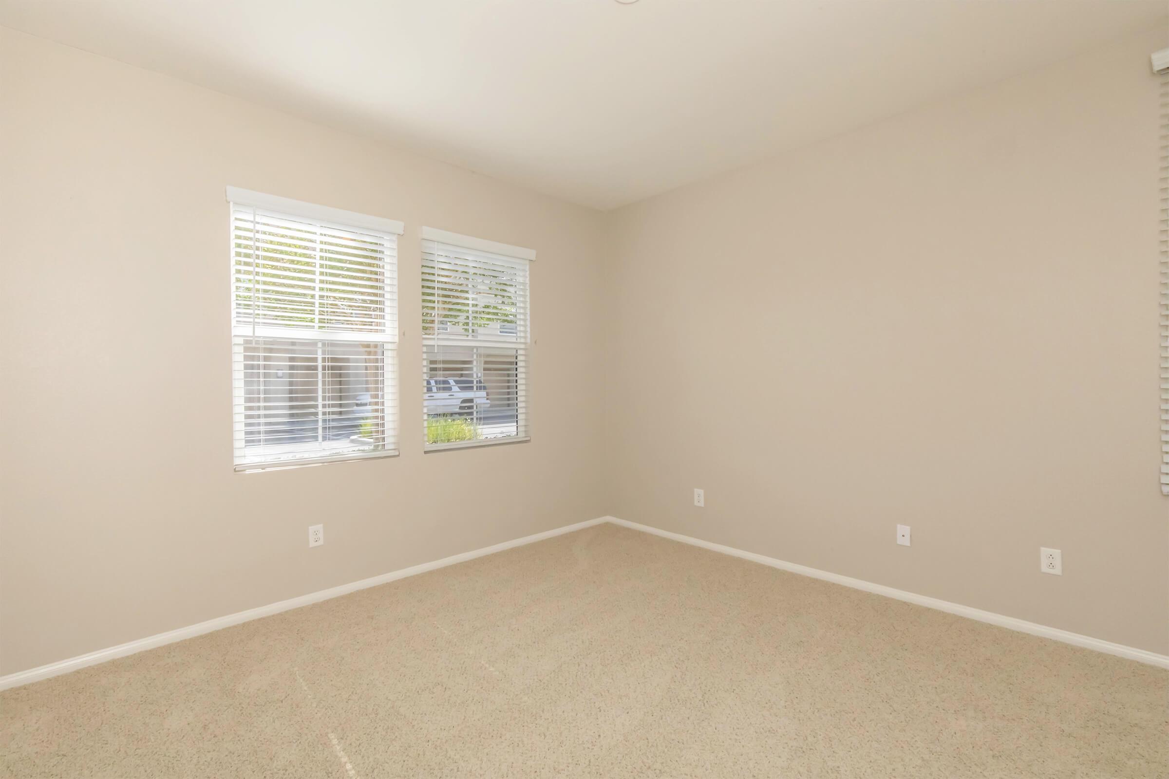 Empty room with beige walls and carpet. Two windows with white blinds allow natural light to come in. The room has no furniture or decorations, creating a spacious and neutral atmosphere.