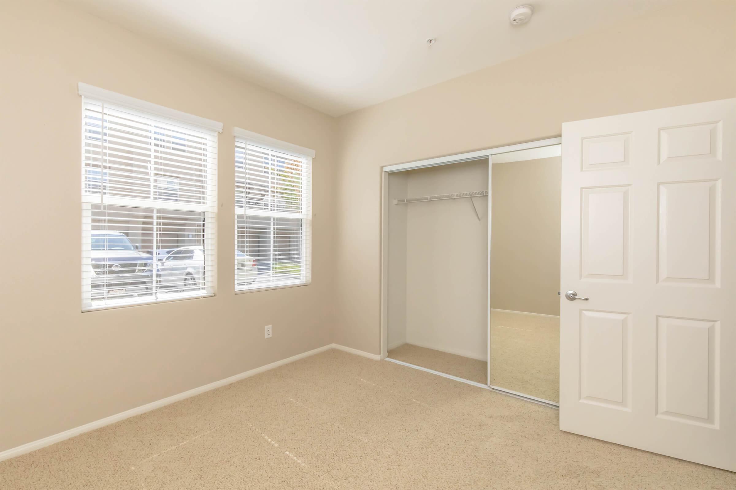 Empty bedroom with beige walls and carpeted floor. Two windows with white blinds allow natural light. A mirrored closet door is visible, and the room features a single white door. The overall atmosphere is bright and neutral, suitable for various decor styles.