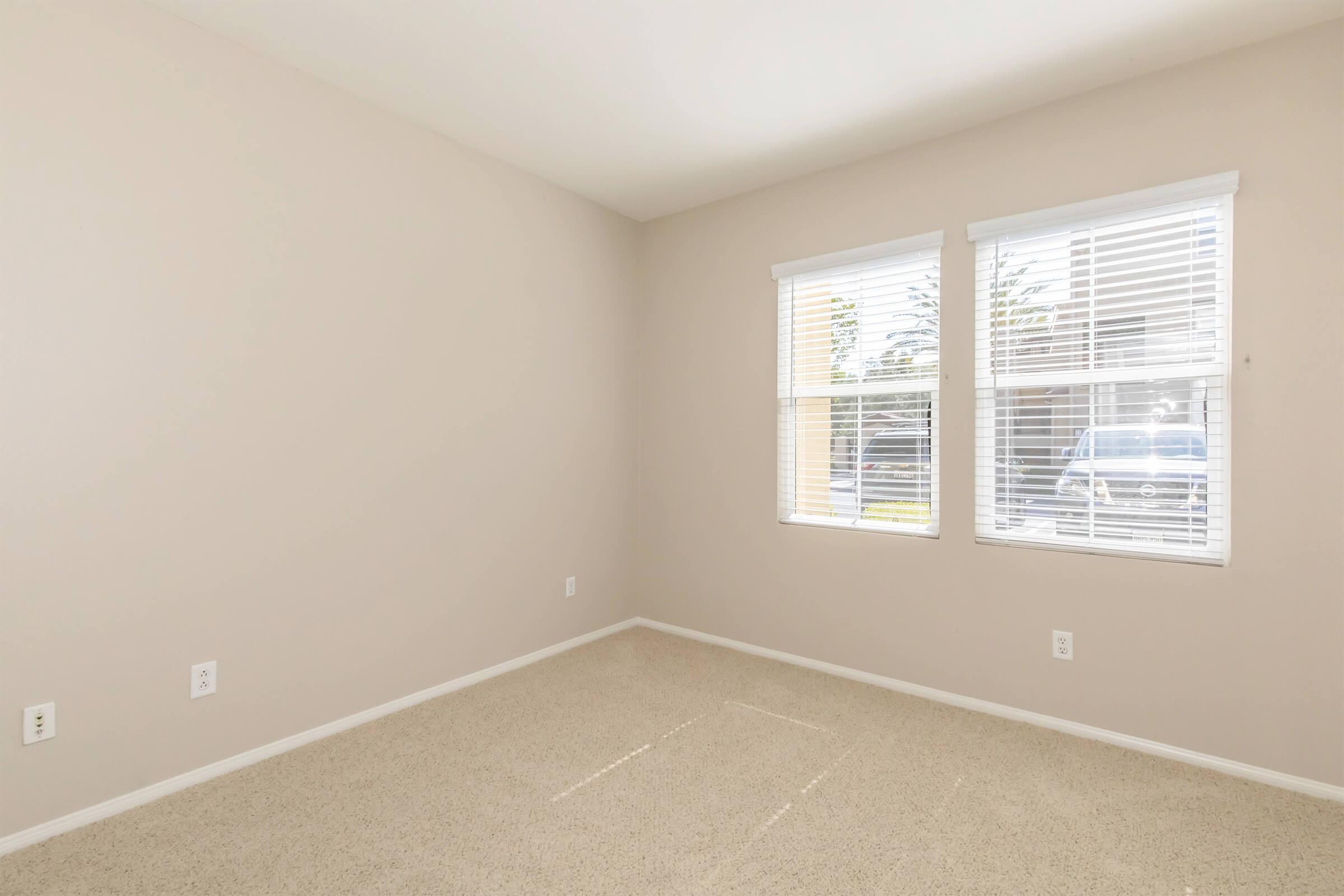 Empty room with beige walls and light beige carpet. There are two windows with white blinds allowing natural light in. The room has no furniture and is well-lit, creating a spacious feel.