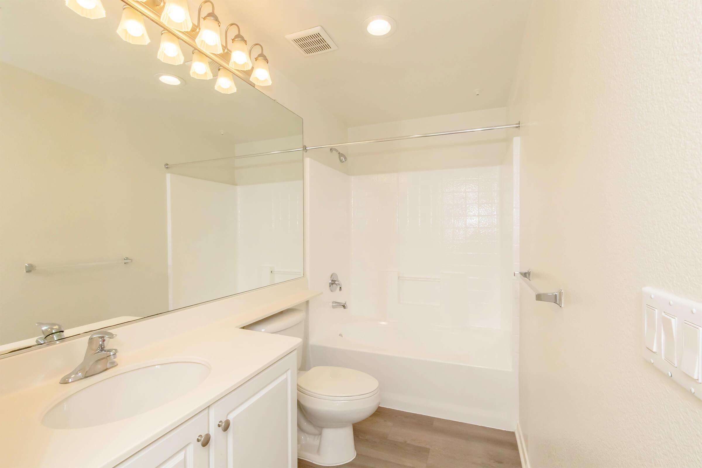 Modern bathroom featuring a white countertop with dual sinks, large mirror, and bright lighting. A white bathtub with shower combination is against the wall, and a toilet is nearby. The walls are painted in a light color, and the floor has a light wood finish. Overall, the space looks clean and well-maintained.