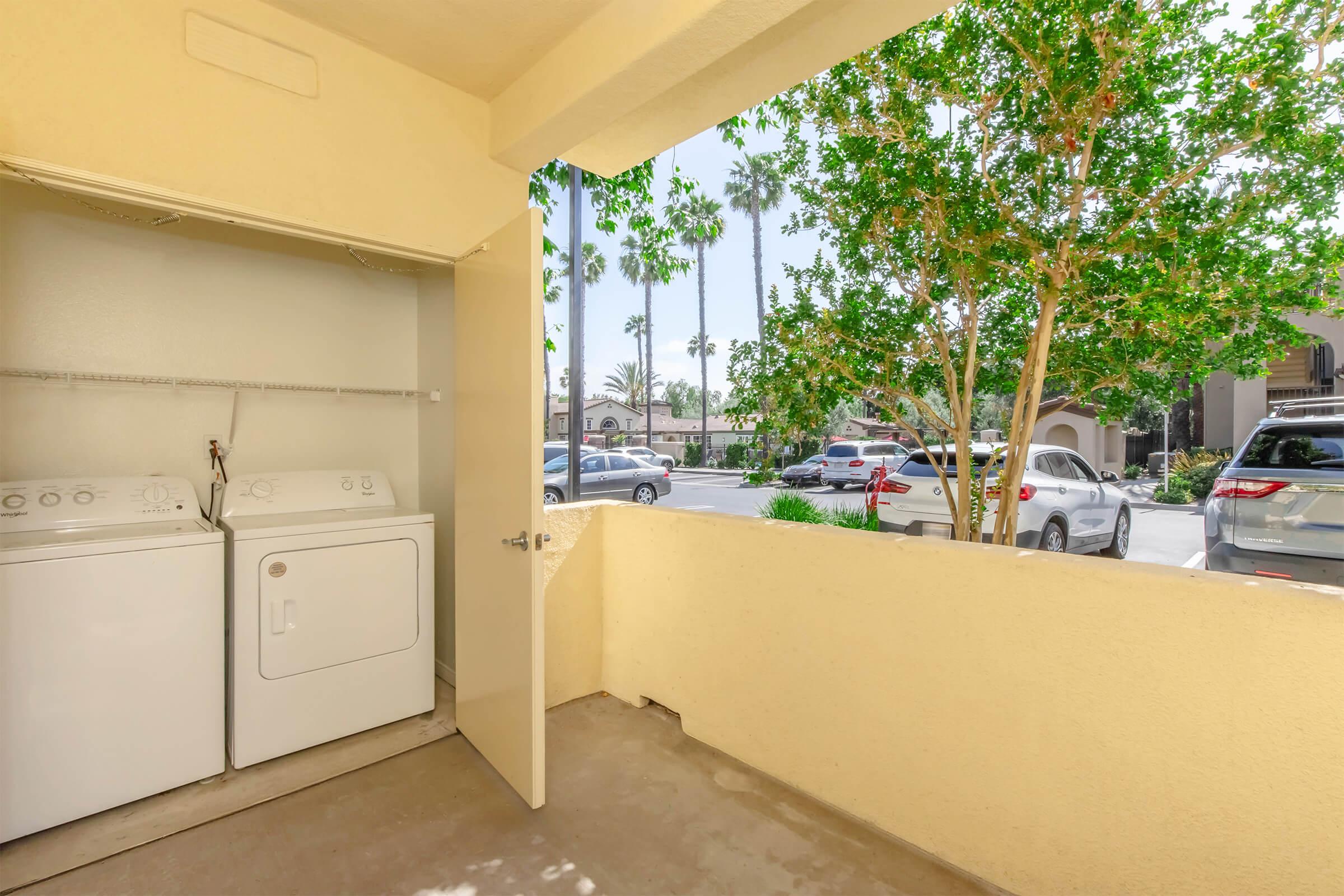 A view from a balcony showing a laundry area with a washing machine and dryer, set against a backdrop of palm trees and parked cars in a residential area. The bright, airy space features beige walls and a wide window, allowing natural light to enter.