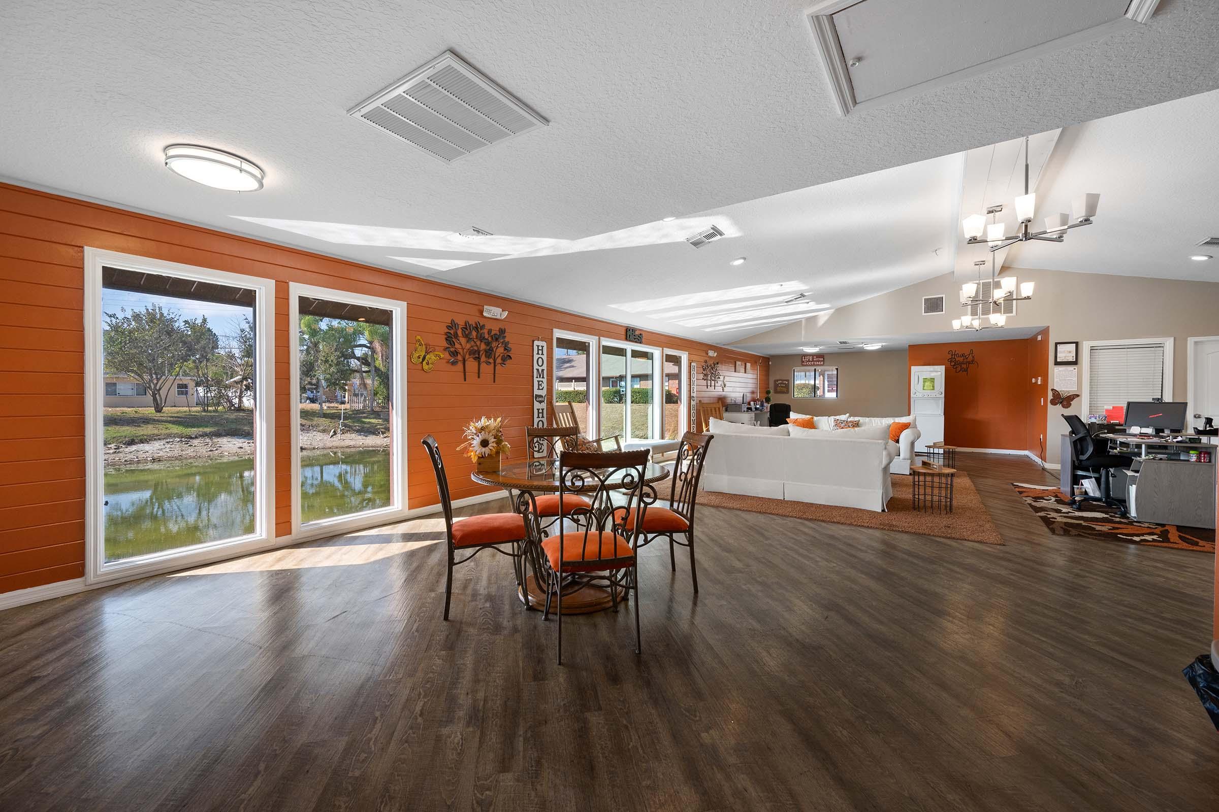 Open concept living area featuring a dining table with orange accents, surrounded by large windows that offer a view of a peaceful outdoor setting. The space includes a cozy living room with a white sofa, modern decor, and a workspace with a desk in the background. Natural light fills the room, enhancing the inviting atmosphere.