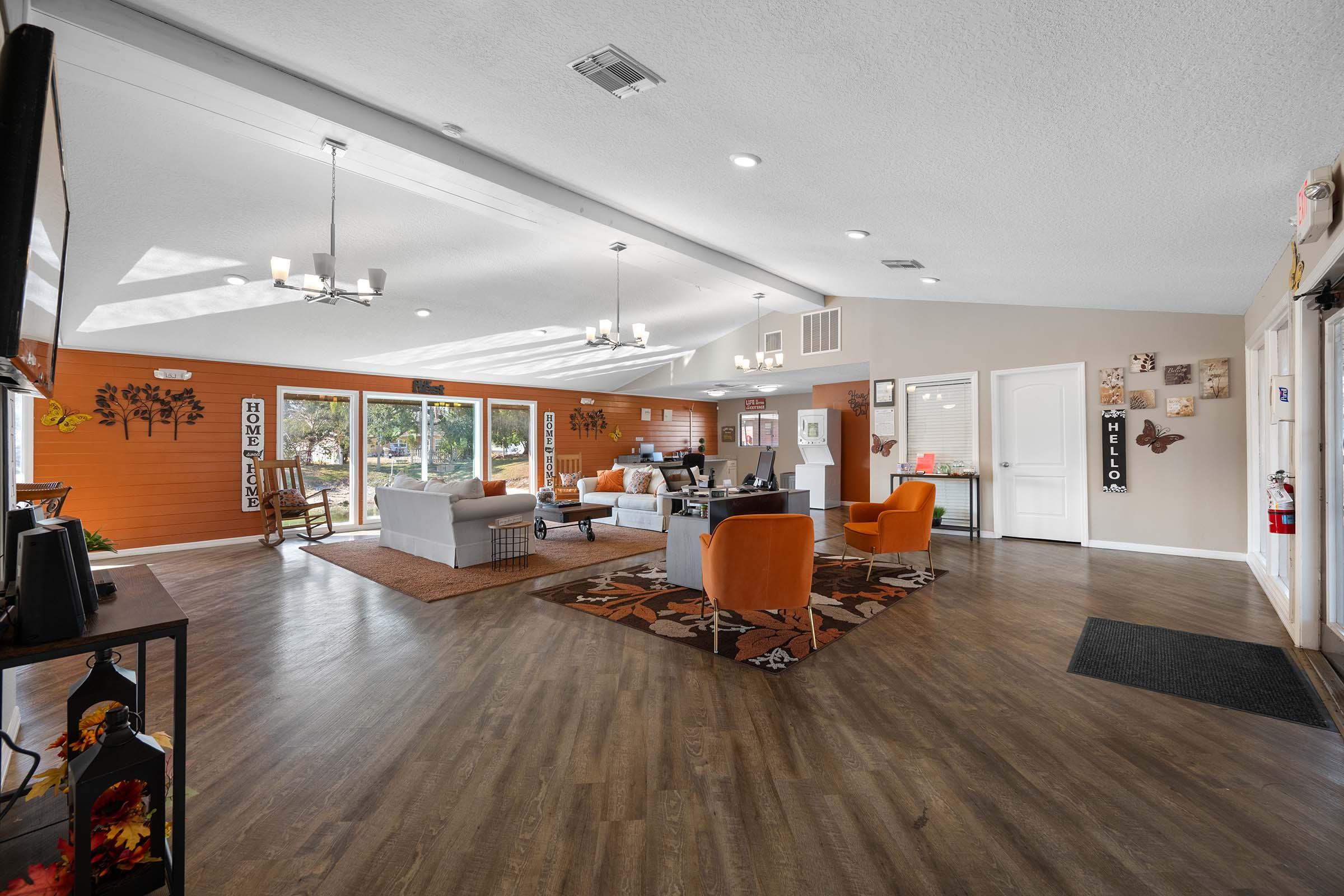 A spacious, modern lounge area with large windows allowing natural light. The room features comfortable seating, including gray sofas and orange chairs, a coffee table, and decorative wall art. A TV is mounted on one wall, and there are plants and decor elements that enhance the welcoming atmosphere.