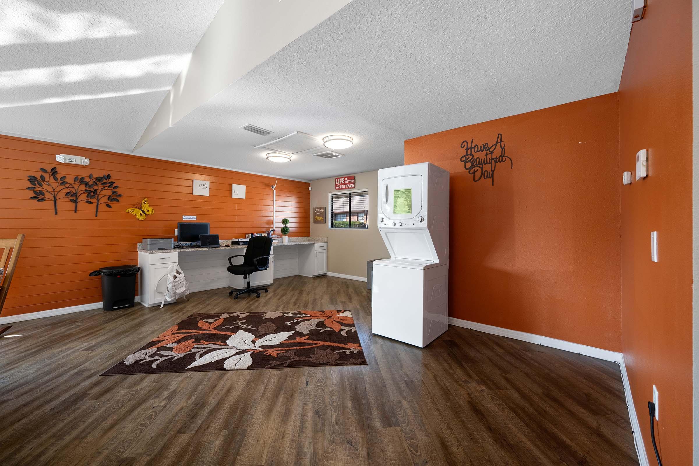 A cozy room featuring an orange accent wall, wooden flooring, a decorative area rug, a laundry machine, a desk with a chair, and wall art. The space is well-lit with natural light streaming in through windows, creating a welcoming atmosphere designed for relaxation or productivity.