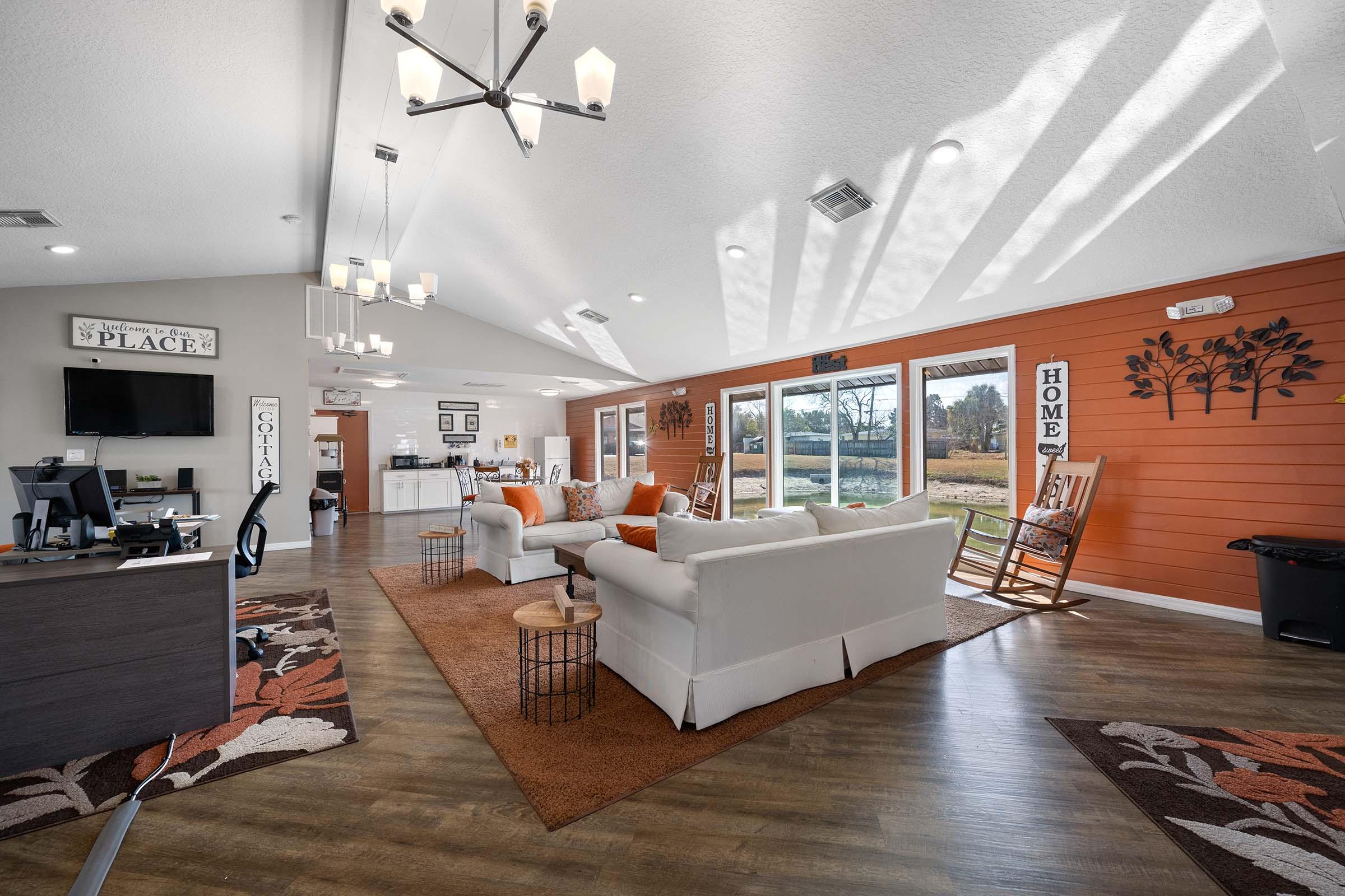 A cozy interior space featuring a bright, open layout with large windows. The room includes a seating area with a white sofa and orange cushions, a decorative rug, wooden rocking chairs, and a reception desk. Sunlight streams in, illuminating the space, which has warm wooden tones and decorative wall art.