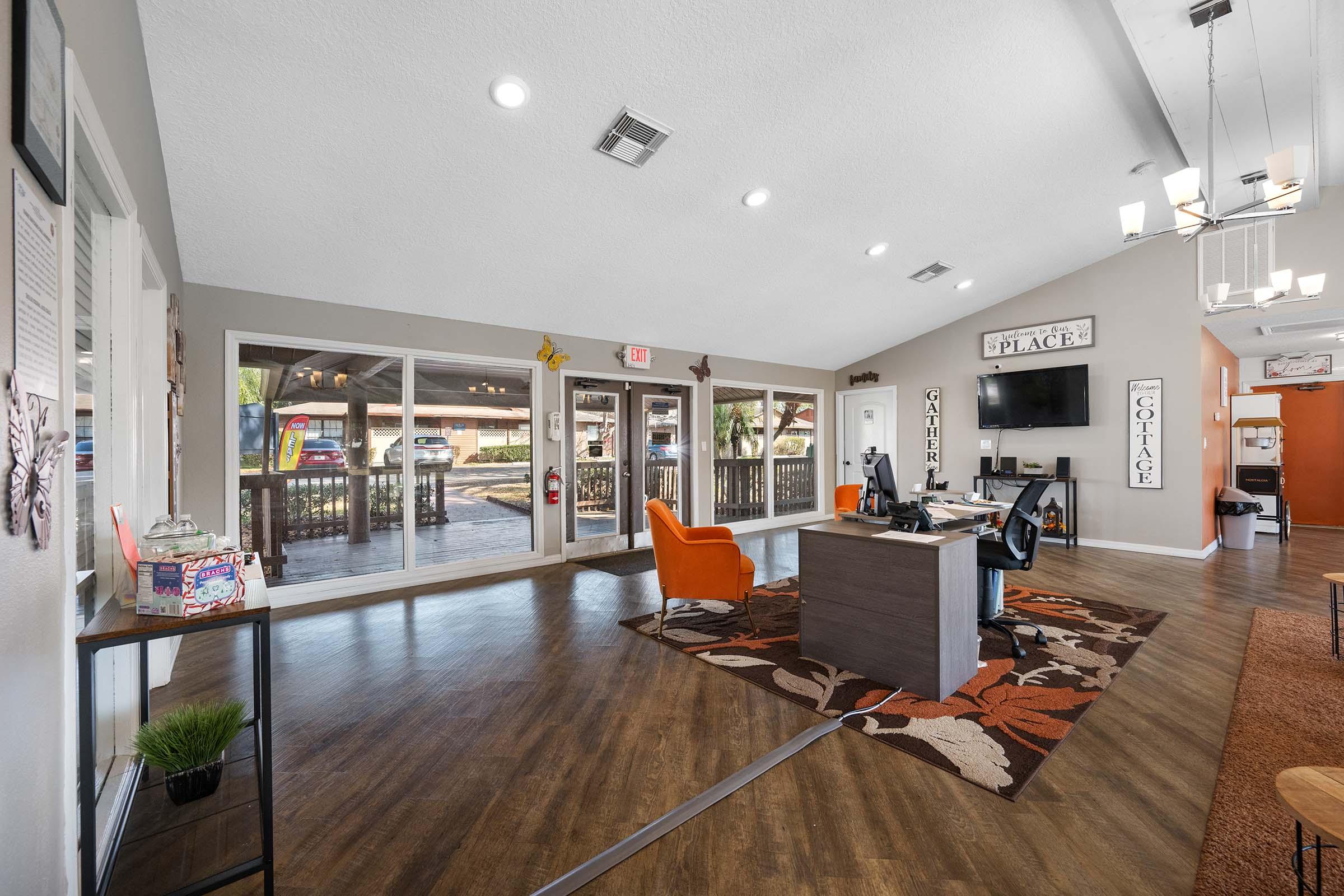 A modern office reception area featuring a wooden desk with a computer, an orange armchair, and decorative wall signs. Large windows allow natural light to illuminate the space, which has a warm color palette and stylish flooring with a floral rug. A welcoming atmosphere is created with plants and decor items.