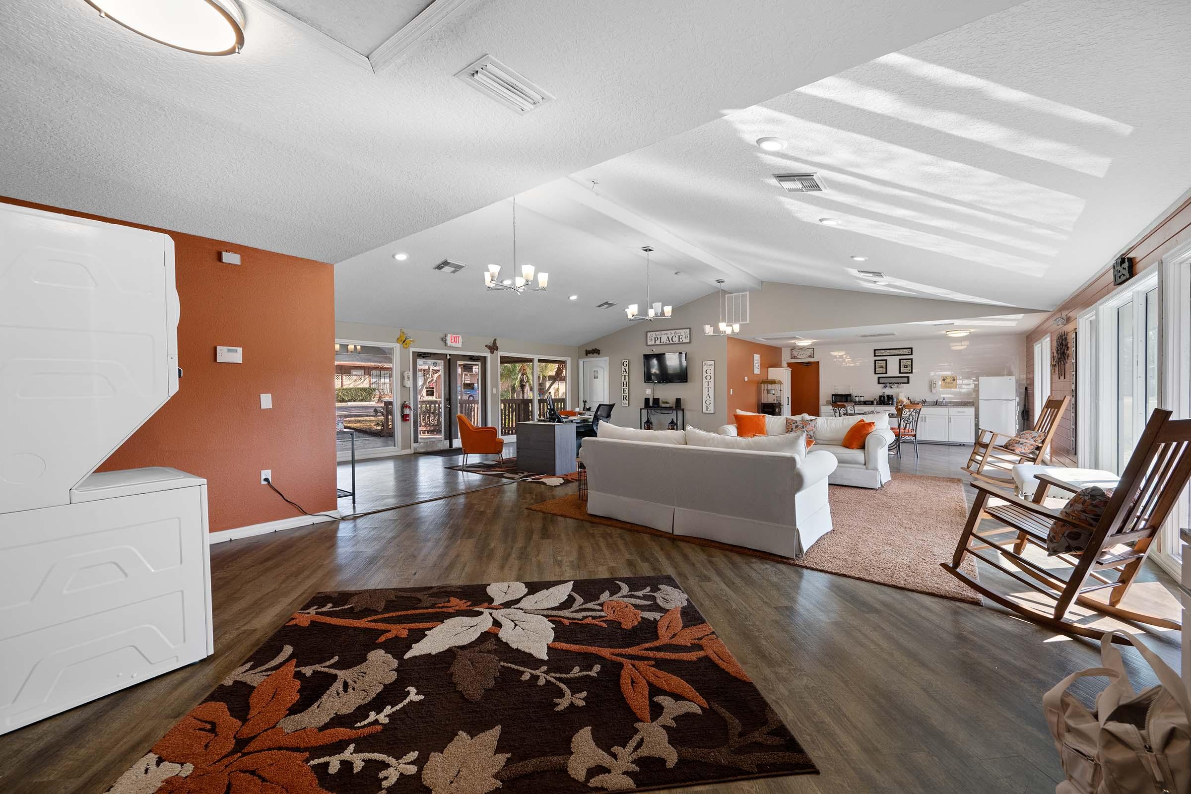 A spacious living area featuring a cozy arrangement with a white sofa and orange accent chairs. The room has large windows allowing natural light and displays a decorative area rug with leaf patterns. In the background, there's a dining space and a modern kitchen. The overall ambiance is warm and inviting.