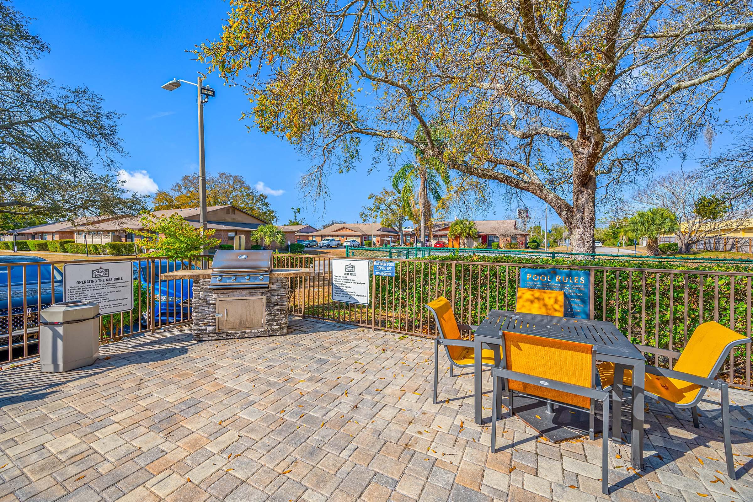 Outdoor area featuring a barbecue grill and a table with chairs, surrounded by a fence. Various trees and plants are visible, and there's a clear blue sky above. In the background, residential buildings and parked cars can be seen, creating a welcoming community space.