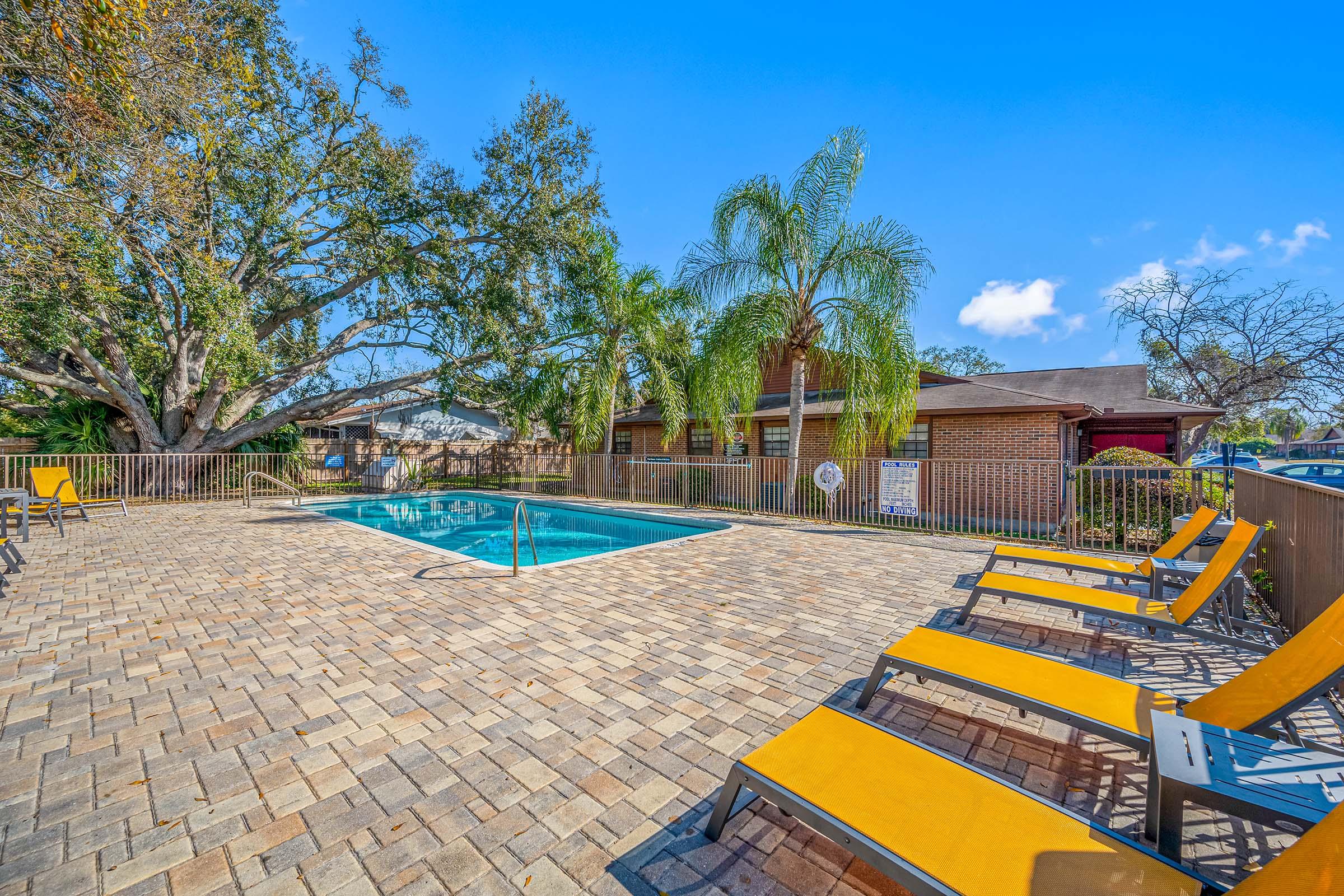 A swimming pool surrounded by a brick patio, decked with yellow lounge chairs. Lush greenery and palm trees provide a tropical atmosphere, while a brick building is visible in the background under a clear blue sky.