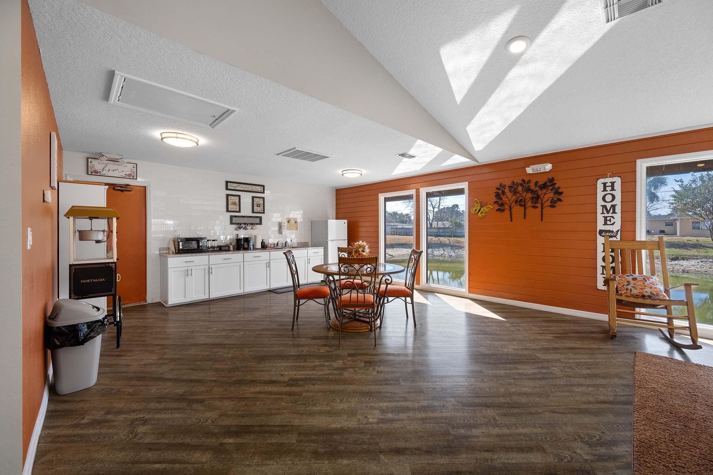 Bright indoor space featuring a dining area with a round table and chairs, a kitchenette with white cabinets, and a window overlooking a pond. The walls are painted orange, with decorative wall art and a warm, inviting atmosphere. A comfortable chair and a trash bin can also be seen in the scene.