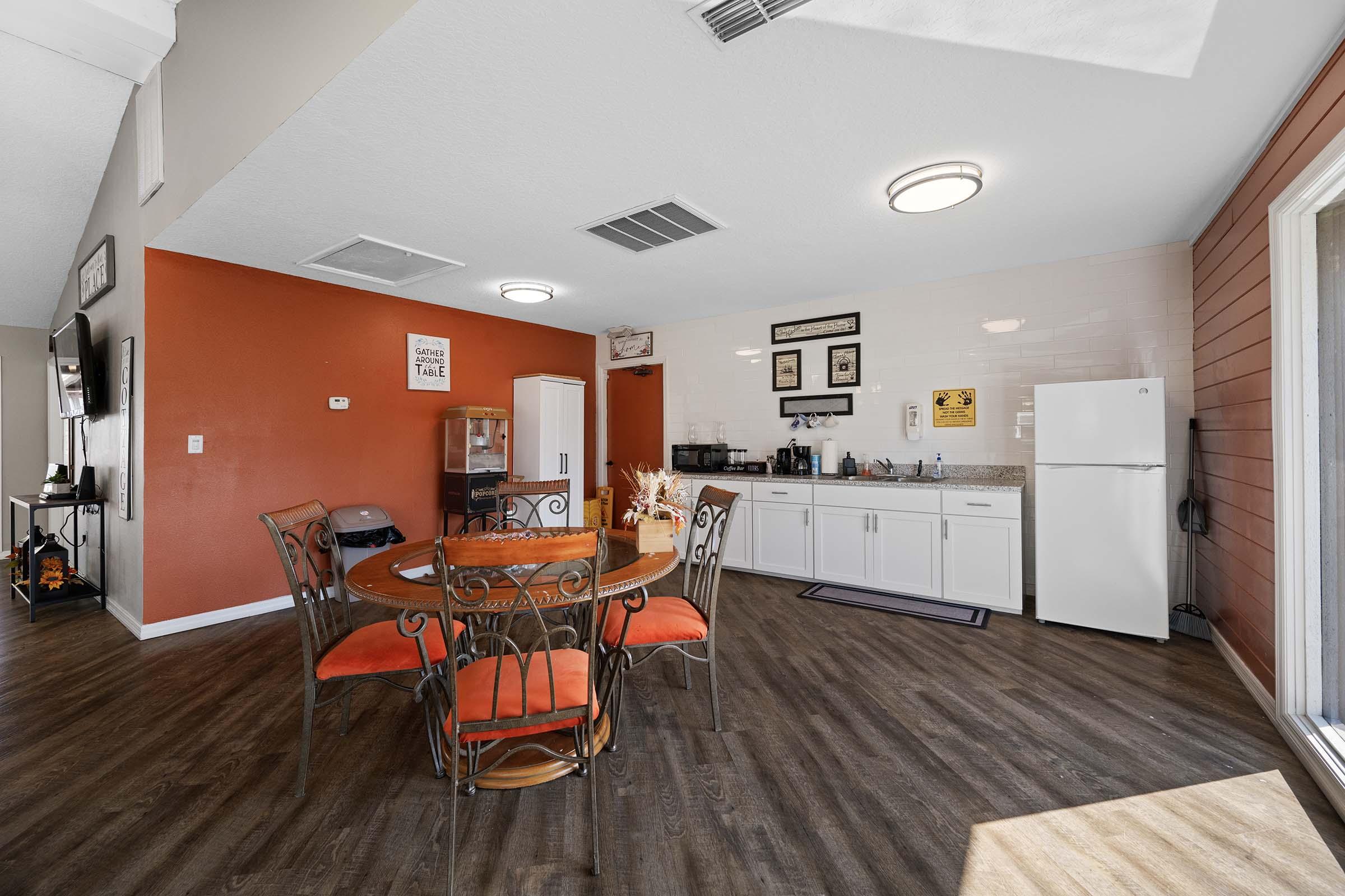 A kitchen area featuring a round dining table with four chairs, orange cushions, and a side kitchen counter. The walls are a mix of orange and white, with a refrigerator, coffee station, and various decorative items. There is a large window letting in natural light, creating a warm and inviting atmosphere.