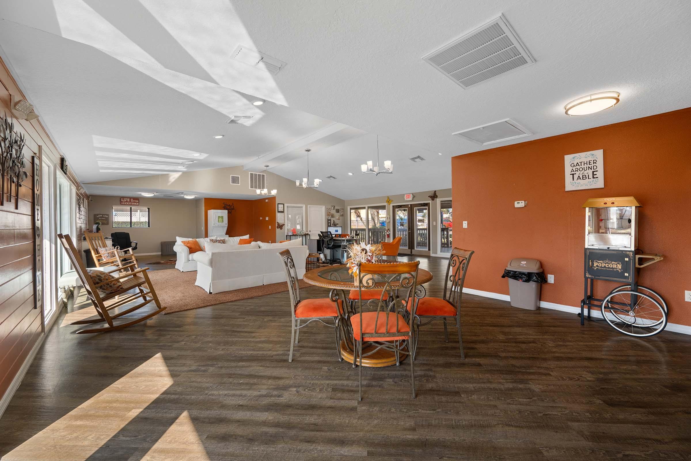 A cozy community lounge area featuring a seating arrangement with a round table and orange chairs, a rocking chair, and a popcorn machine. The walls are painted in warm tones, with natural light streaming in from large windows, creating an inviting atmosphere for social gatherings.