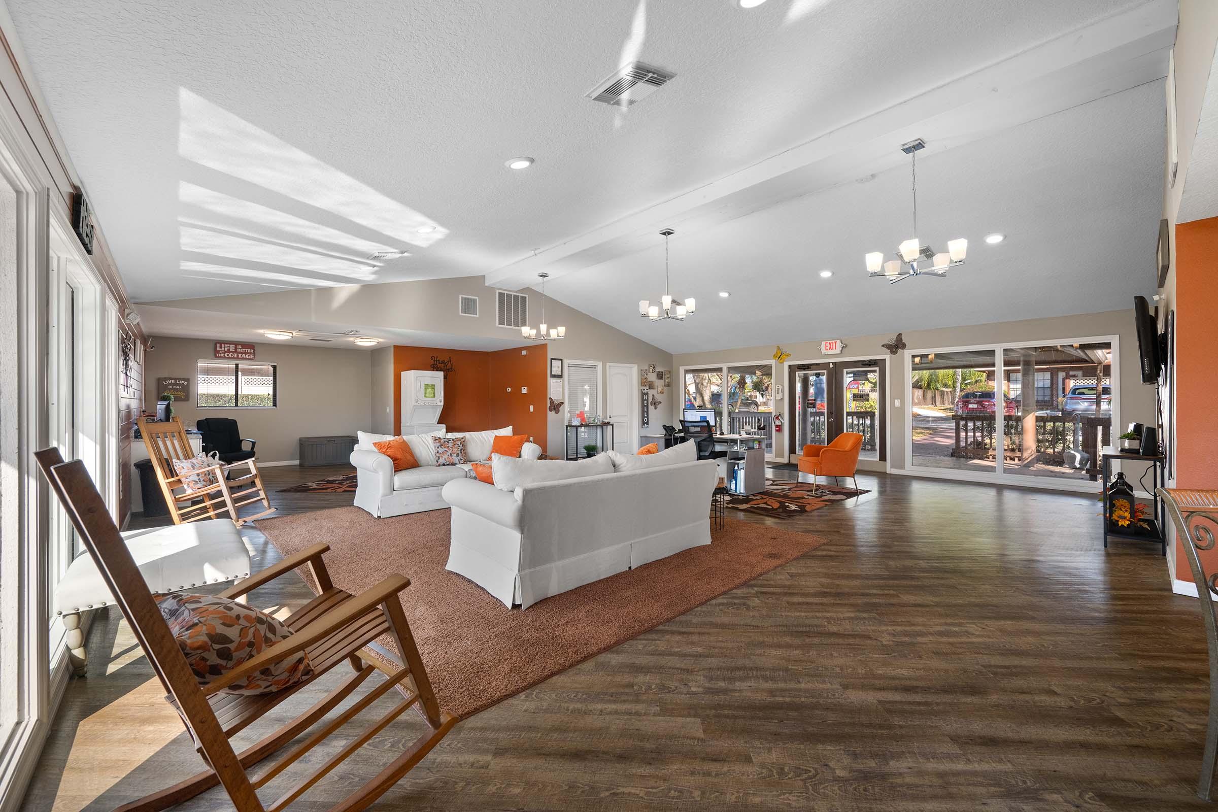 Interior view of a spacious and welcoming lounge area featuring comfortable seating with white sofas and orange accents. Natural light floods the space through large windows. There's a rustic rocking chair, a coffee table, and a carpeted floor. The decor includes warm colors and a casual atmosphere, ideal for relaxation or socializing.