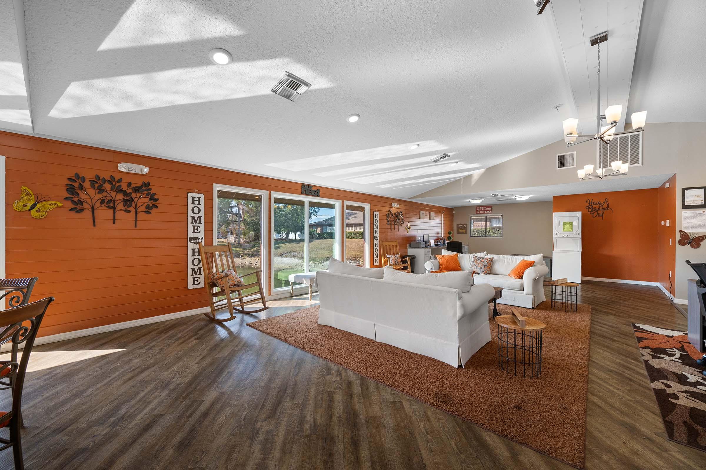 Cozy living room with an orange accent wall, featuring a white sofa adorned with orange pillows, a rustic rocking chair, and a textured area rug. Large windows allow natural light, creating a warm atmosphere. Decorative elements include wall art and a chandelier, enhancing the inviting ambiance.