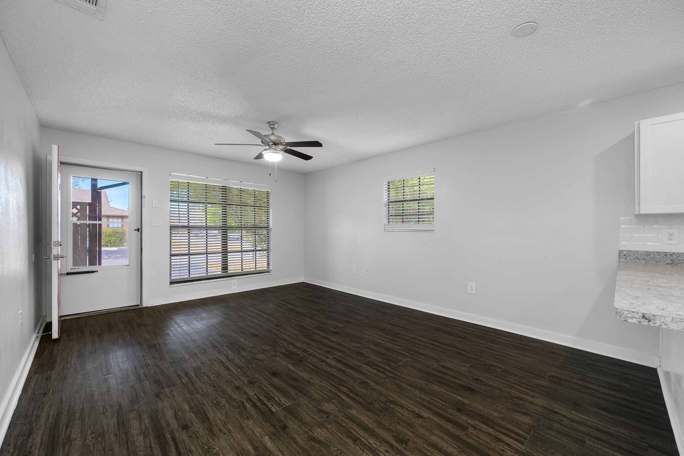 A spacious living area with a ceiling fan, featuring a doorway leading outside and large windows with blinds. The floor is dark wood, and the walls are painted light gray. A kitchenette is visible in the background with a counterspace. Natural light brightens the room.