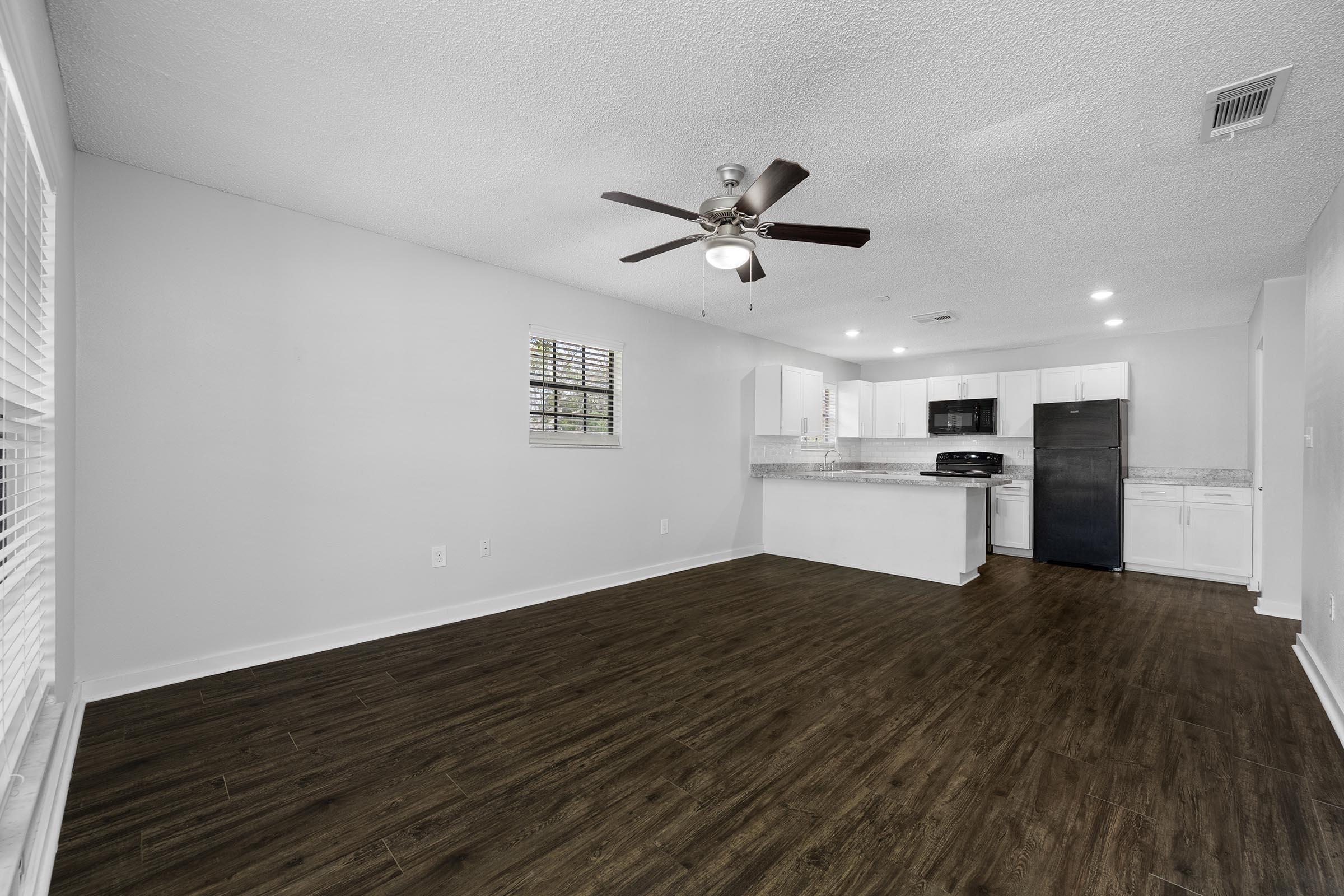 A spacious, modern kitchen and living area featuring dark hardwood flooring, a ceiling fan, white cabinetry, and stainless steel appliances. Natural light enters through a small window with blinds. The layout is open, creating a seamless flow between the kitchen and living space.