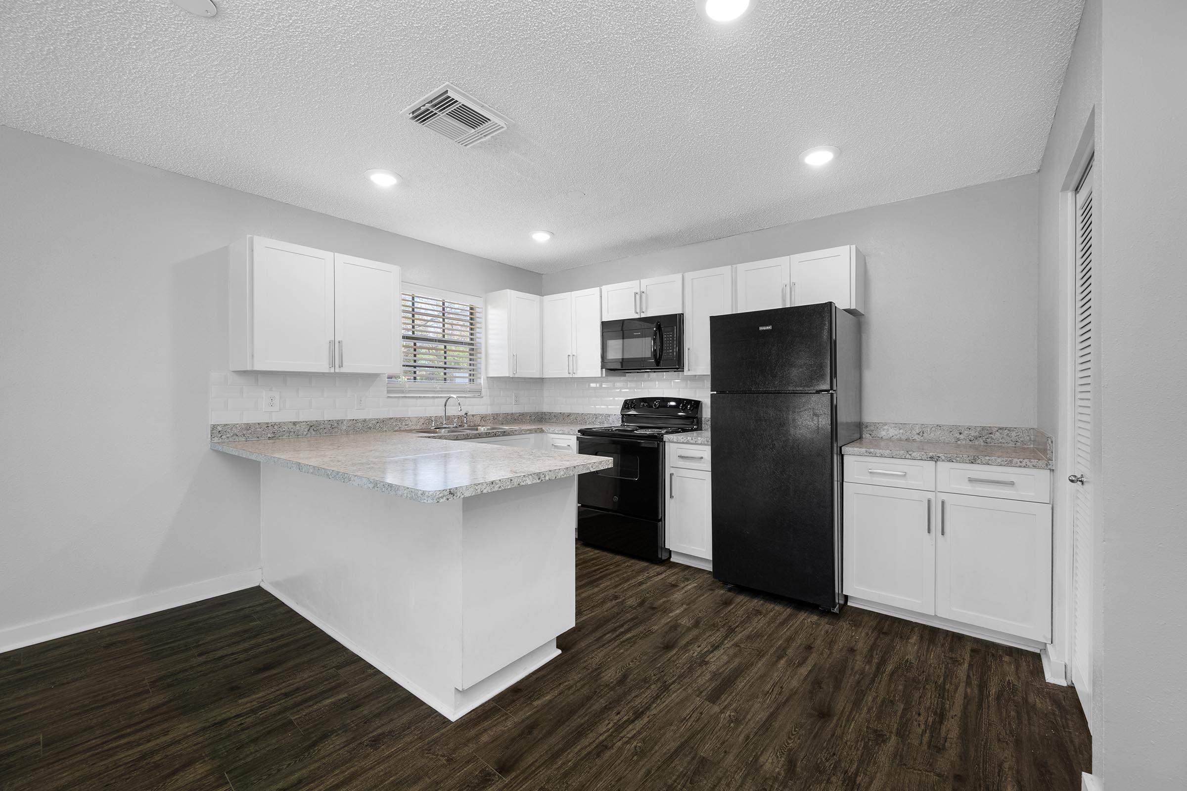 A modern kitchen featuring white cabinetry, a granite countertop island, and stainless steel appliances. The space includes a black refrigerator and stove, with ample natural light coming through a window, and a wood-style floor.