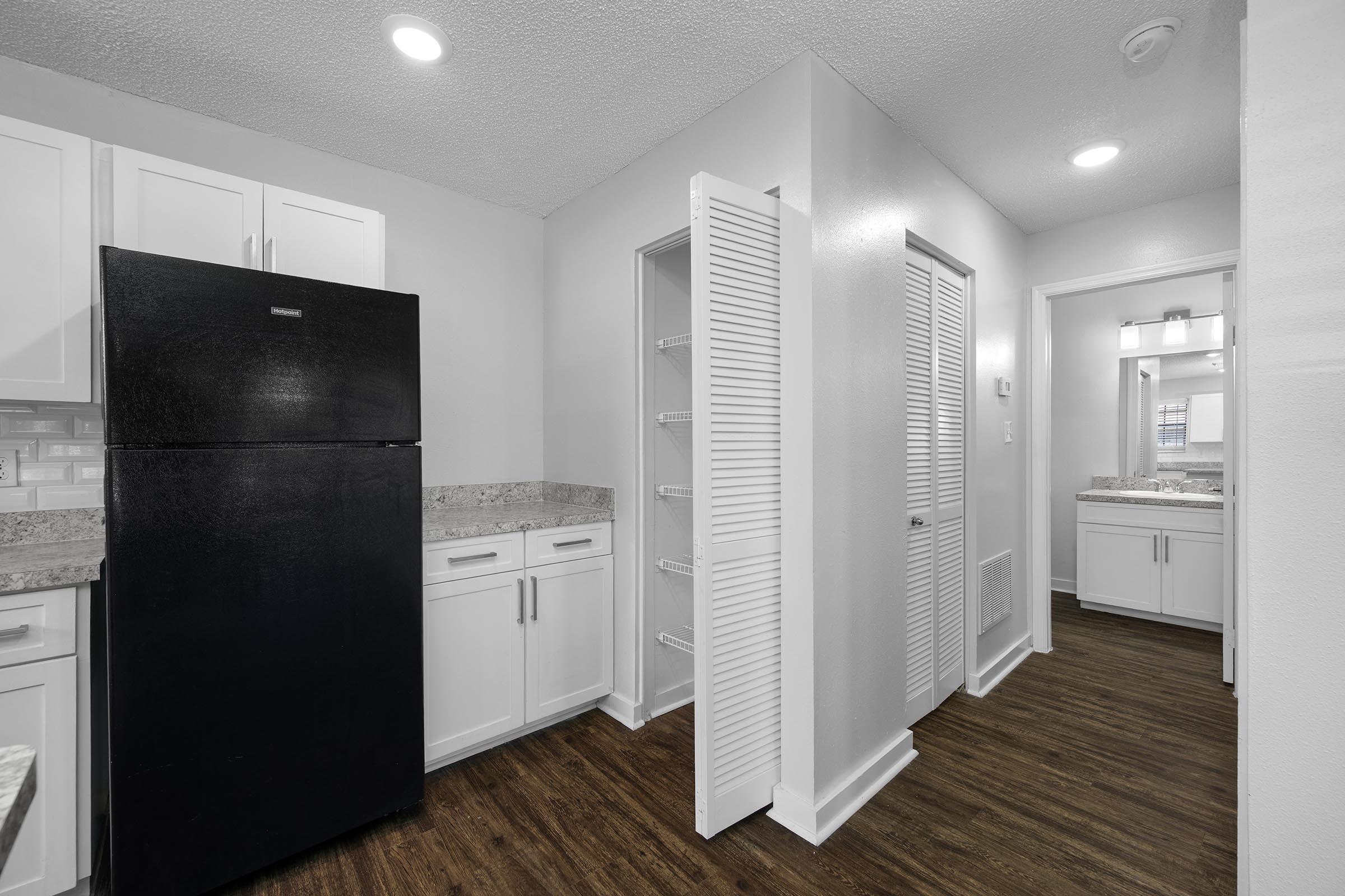 Interior view of a modern kitchen and hallway. The kitchen features white cabinets, a black refrigerator, and granite countertops. A door on the right leads to a bathroom area, with a mirror and light fixtures visible. The flooring is brown, adding warmth to the space. Bright overhead lighting enhances the room's ambiance.
