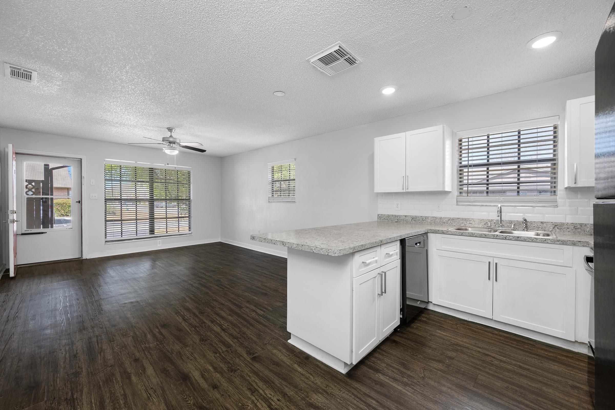 A modern kitchen and living area featuring white cabinets, granite countertops, and stainless steel appliances. Large windows provide natural light, and the space has dark wood flooring. A ceiling fan is visible, and there is an open layout connecting the kitchen to the living area.