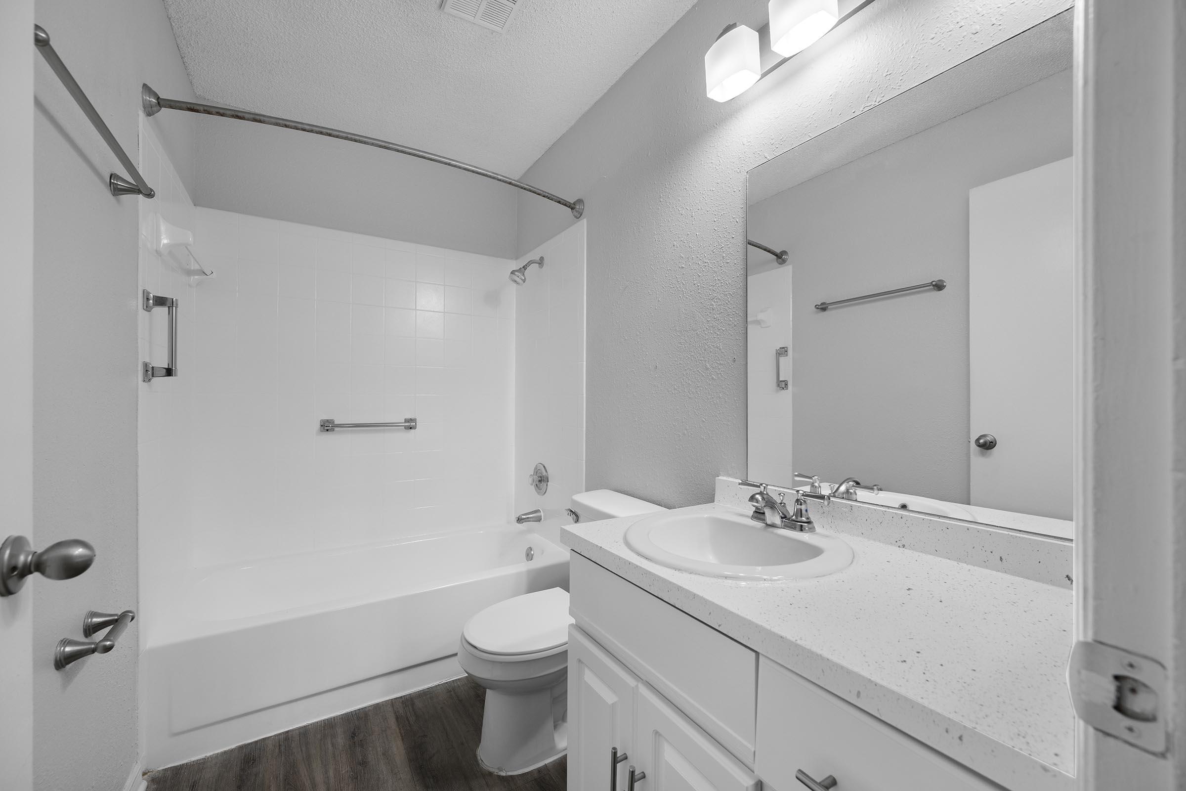 A modern bathroom featuring a white bathtub with a curved shower rod, a single vanity with a sink, and a large mirror. The walls are light gray, and the flooring is dark laminate wood. Bright overhead lights provide ample illumination.