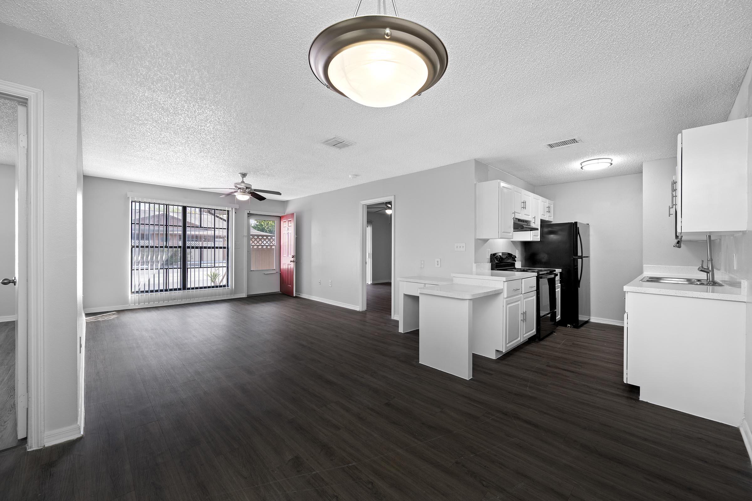 A bright and open living space featuring a modern kitchen with black appliances, a dining area, and a ceiling fan. The room has light-colored walls and laminate flooring, with large windows allowing natural light and a view of the outdoors. The layout is spacious and inviting.