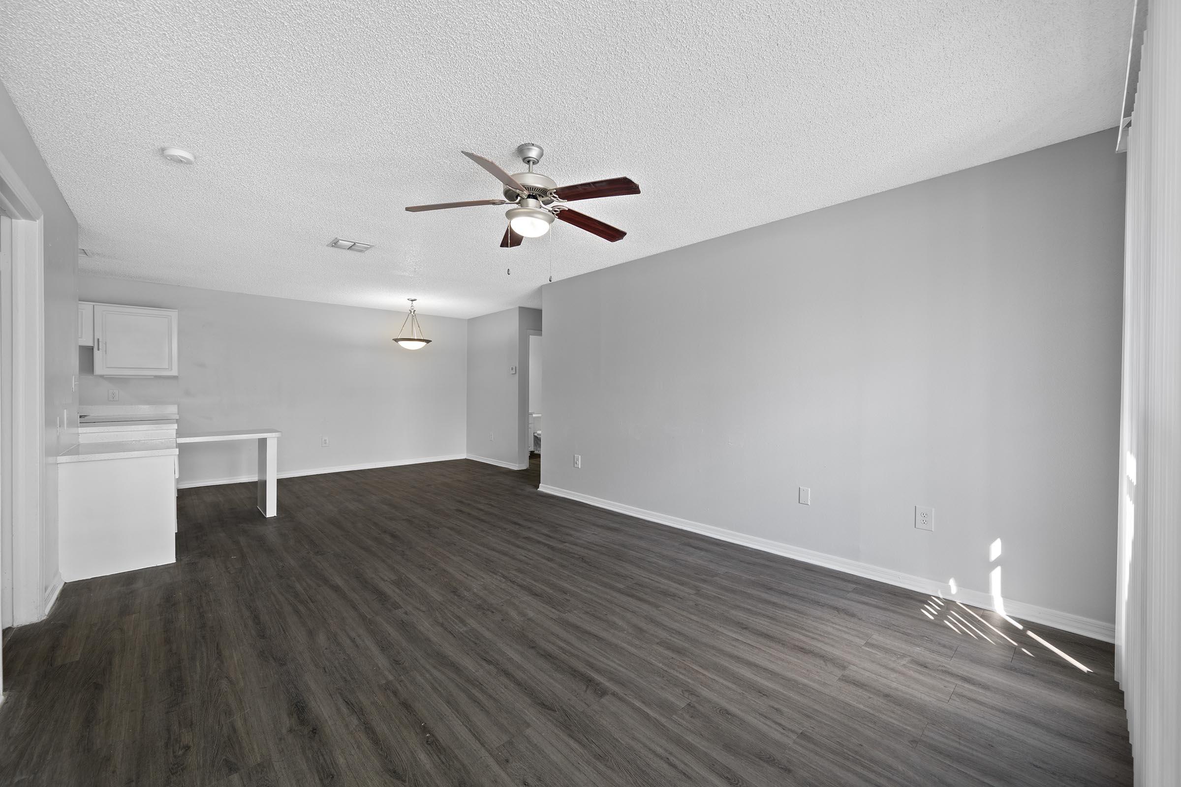 Spacious interior of a modern apartment featuring a ceiling fan, light-colored walls, and dark wood flooring. There's an open area with a small dining space, and a kitchen visible in the background. Natural light streams in from the right side, enhancing the airy feel of the room.