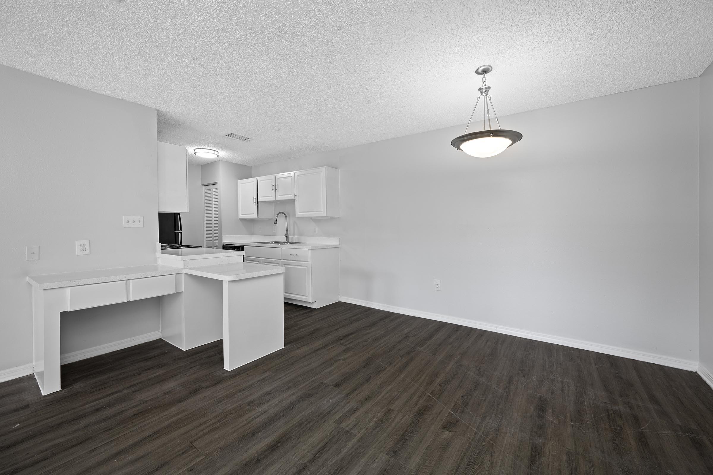 A spacious and minimalist interior of an apartment featuring a kitchen area with white cabinetry and a small desk space. The floor is dark wood, and the walls are painted a light color. A ceiling light fixture hangs in the center, illuminating the open living area.