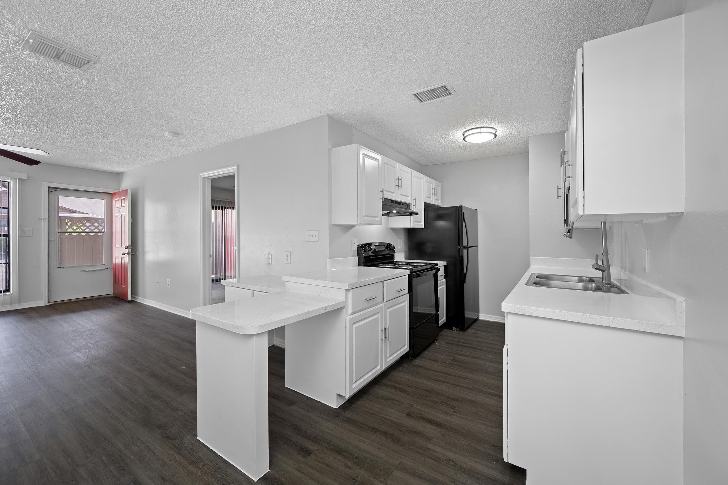 Modern kitchen with white cabinets, black appliances including a refrigerator and oven, and a countertop island. The space features laminate flooring and a bright, airy atmosphere with natural light coming through a nearby window. An open layout connects the kitchen to a living area with a door leading outside.