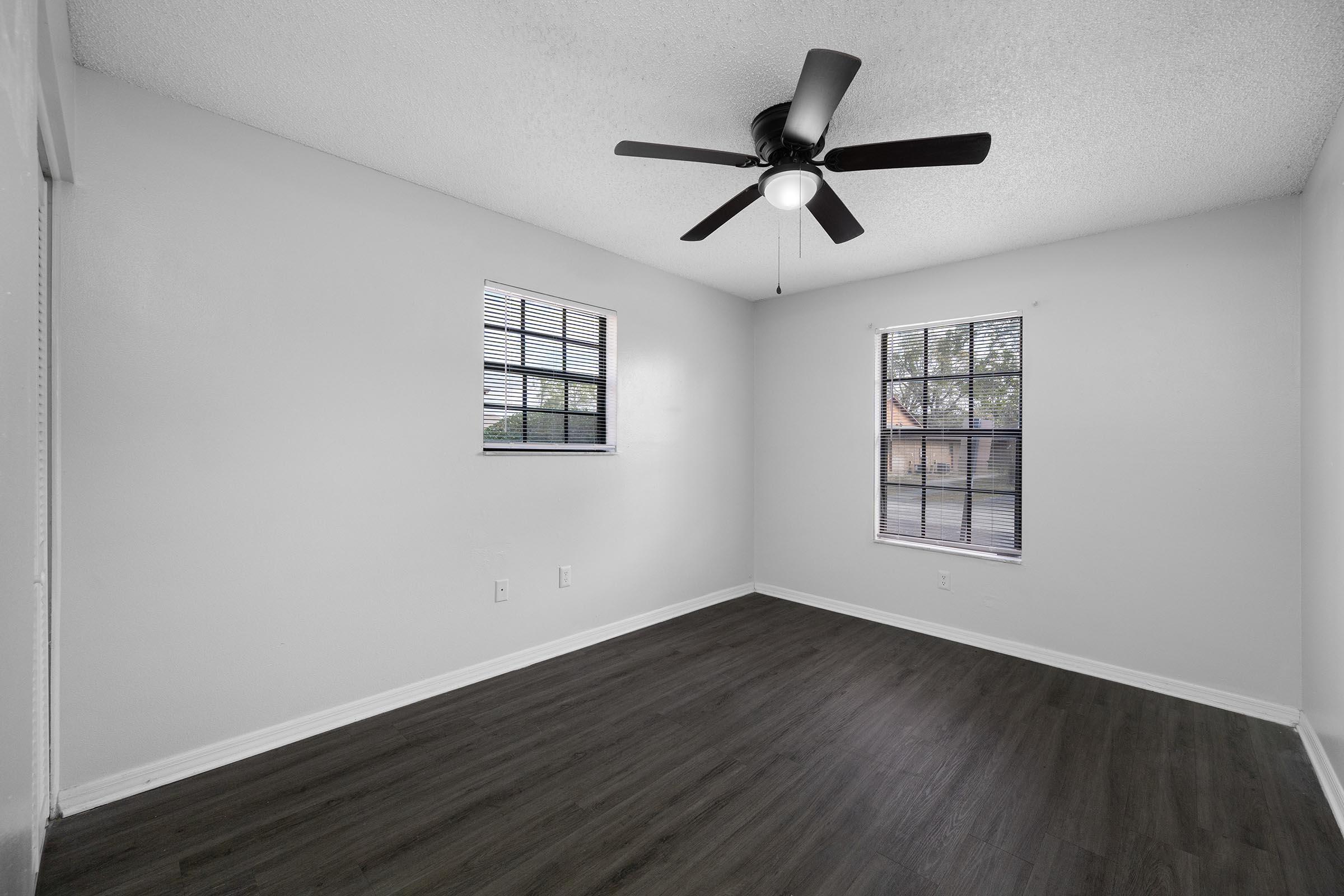 Empty room with light gray walls and dark wood-like flooring. Features include a ceiling fan with a light fixture and two windows with black blinds, allowing natural light to enter. The room is uncluttered, creating a spacious appearance.