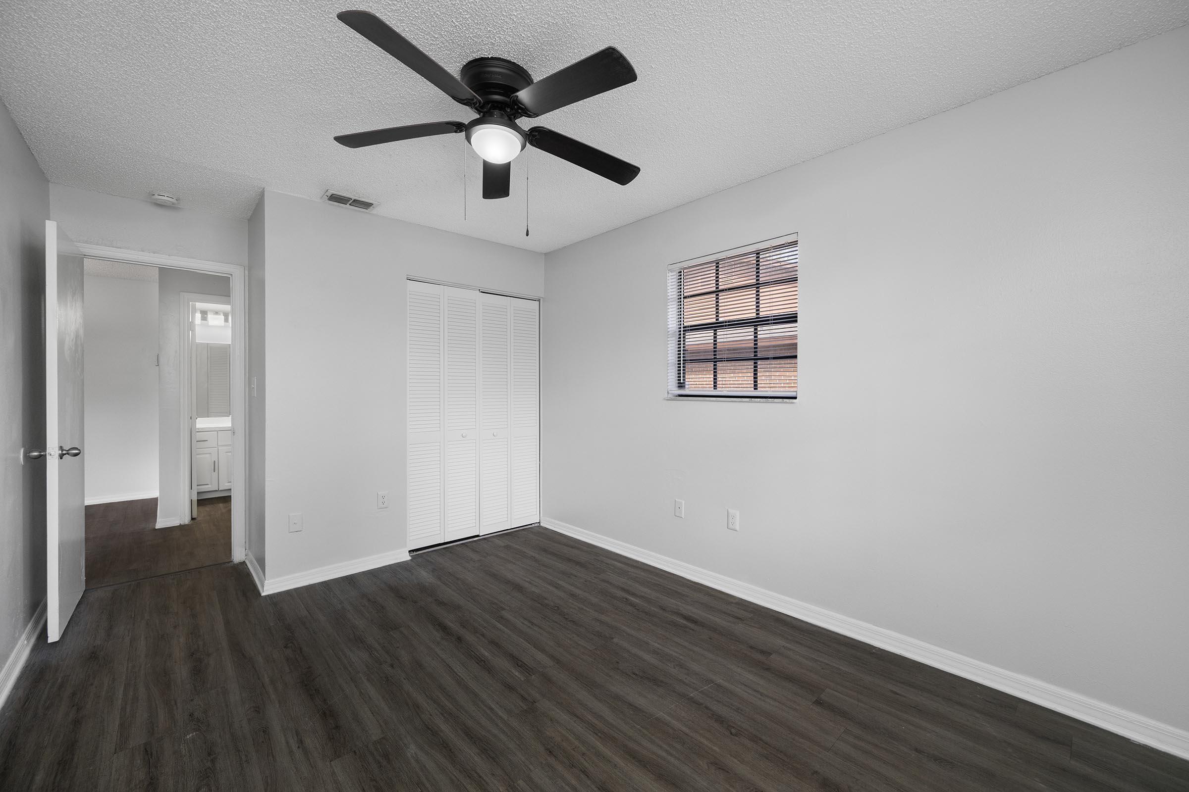 A minimalist, empty room with light gray walls and a ceiling fan. It features a window with blinds, a closet with sliding doors, and a doorway leading to another room. The flooring is dark wood, creating a modern and clean aesthetic.