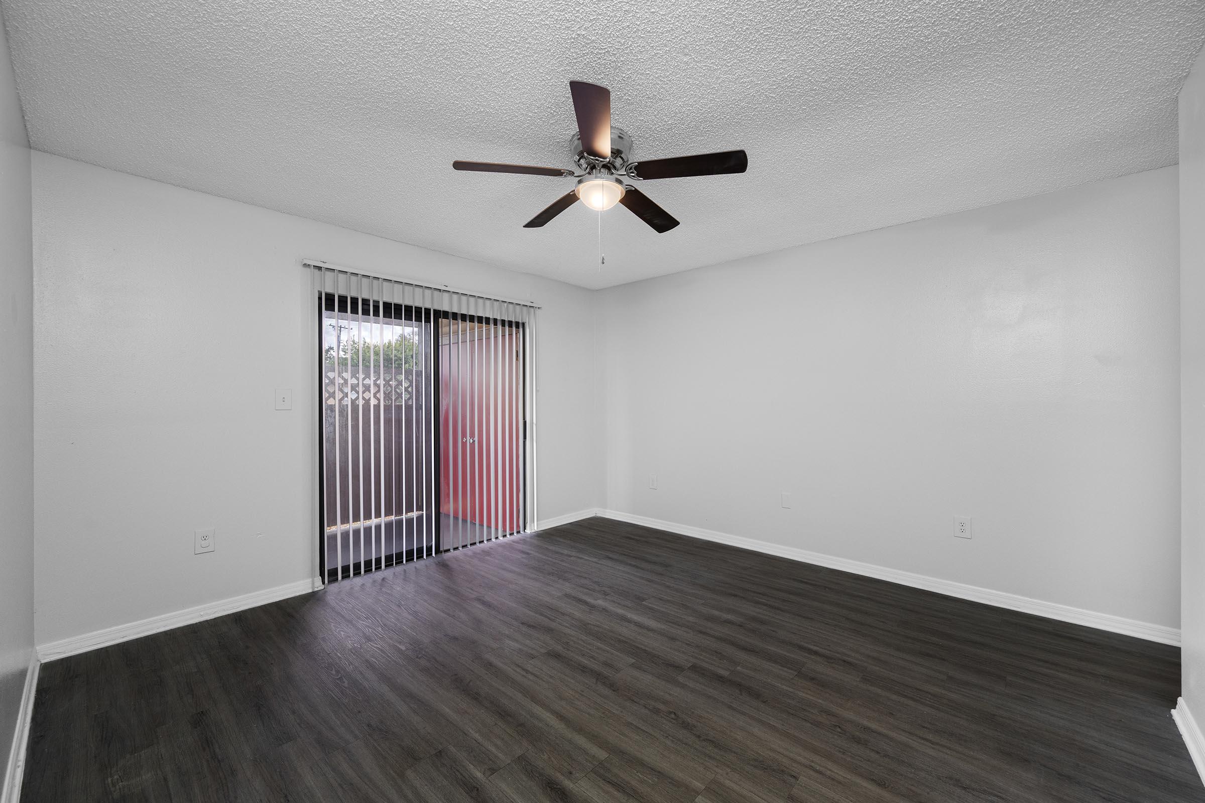 A spacious room with a ceiling fan and wooden flooring. The walls are painted white, and there are vertical blinds covering a sliding glass door that leads outside. The room is well-lit, and the overall design is modern and minimalistic.