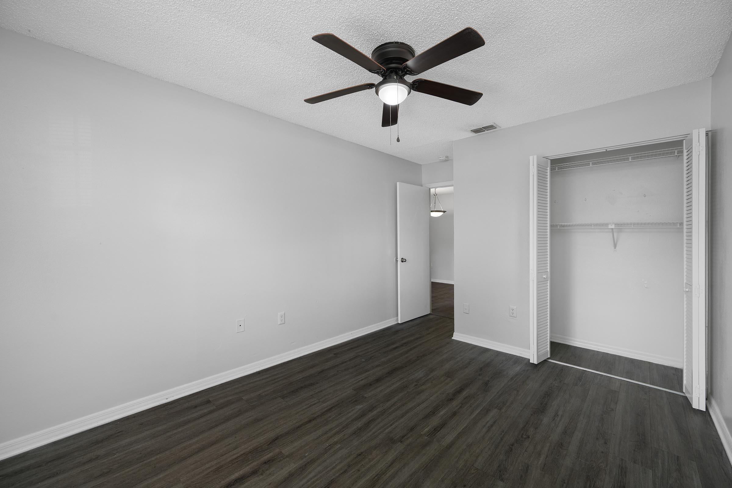 A bright, empty room featuring a ceiling fan, light-colored walls, and dark wood flooring. There is a closed door on the left and a closet with sliding doors on the right. The space appears clean and well-lit, with a view into another room beyond the doorway.
