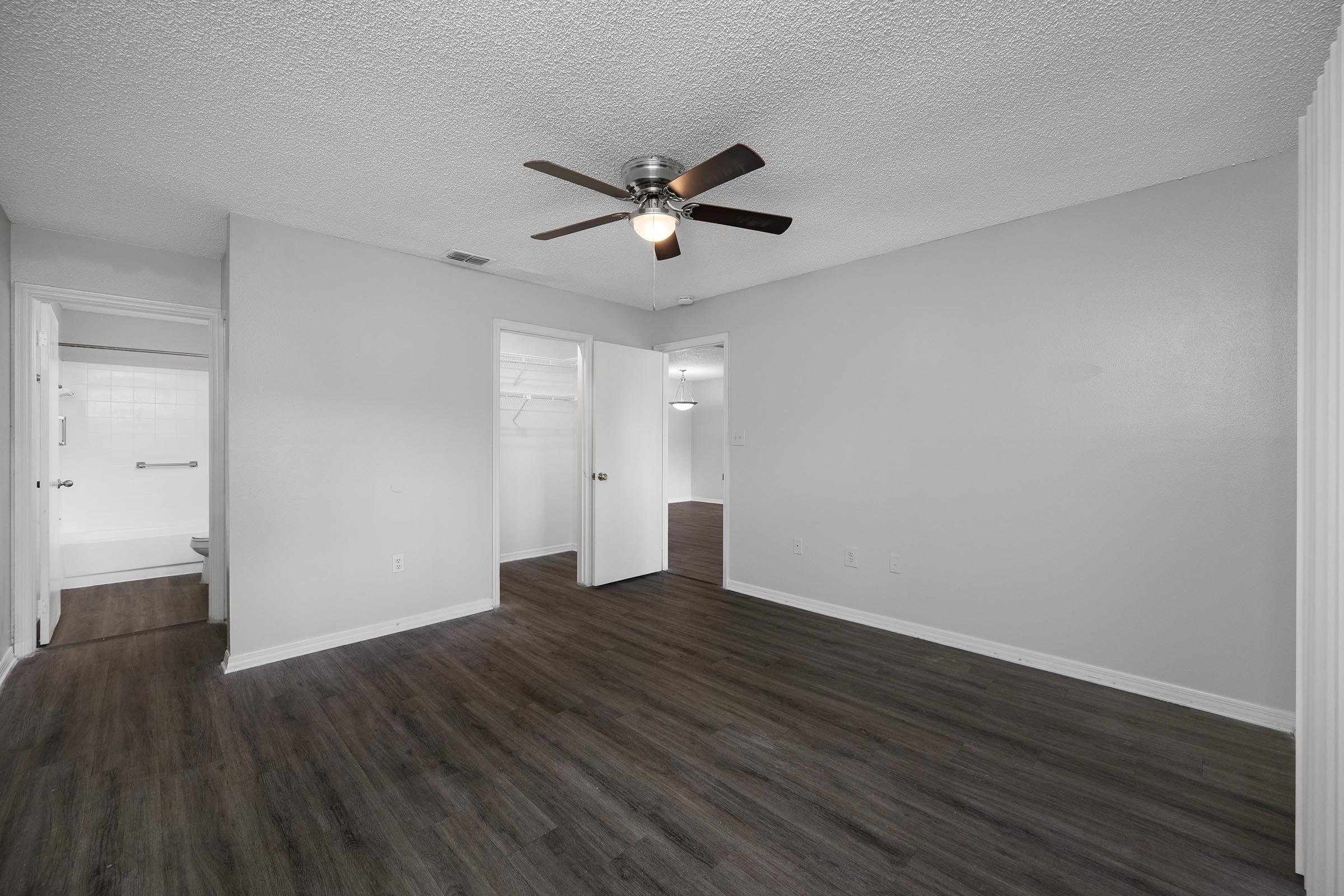 A spacious and well-lit interior of an apartment, featuring a ceiling fan, light gray walls, and dark wood flooring. Two doorways lead to other rooms, with a closet visible. The overall atmosphere is clean and modern, suggesting a comfortable living space.