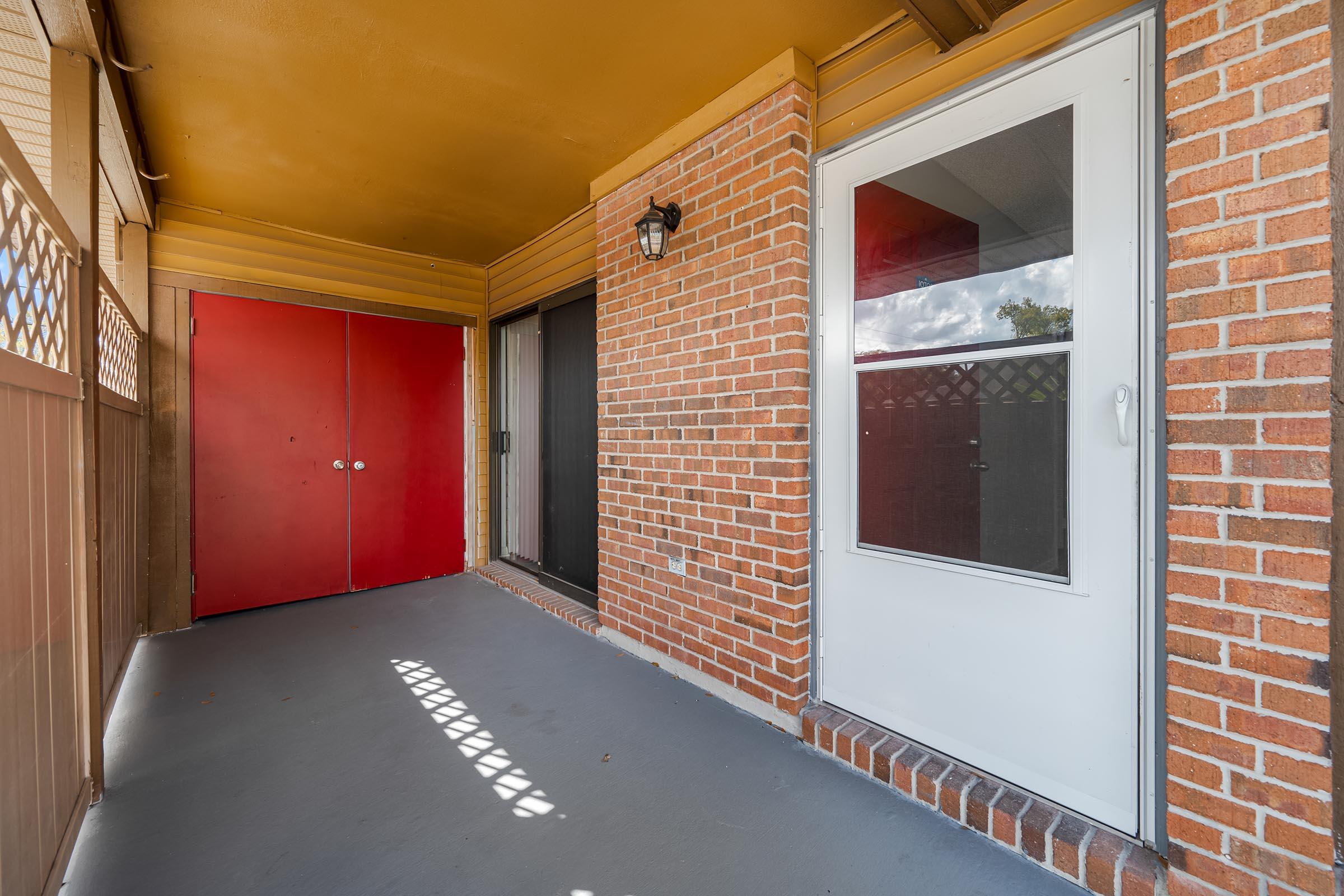 A covered porch area featuring a tan floor, exposed brick walls, and bright yellow ceiling. There are two doors: a solid red double door on the left and a white single door on the right, with a light fixture mounted beside the white door. Sunlight casts shadows on the porch floor.