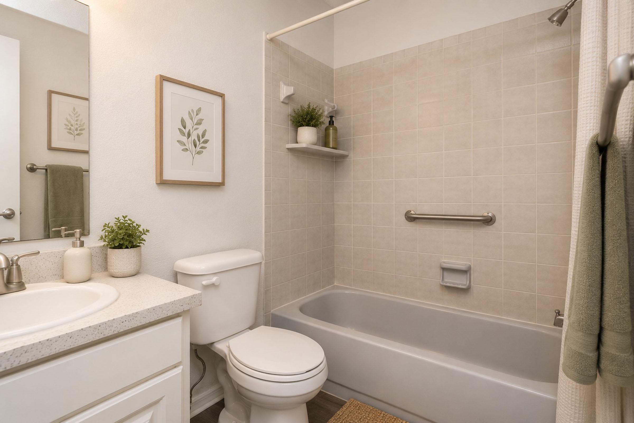 A clean and modern bathroom featuring a white sink with a mirror above, a toilet, and a bathtub with a shower. Light beige tiles on the walls and a small shelf with plants add a touch of greenery. A framed botanical print is hung on the wall, enhancing the fresh, minimalistic aesthetic.