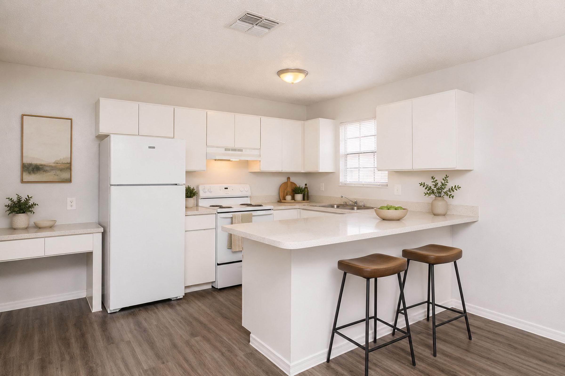 A modern kitchen featuring white cabinetry, a refrigerator, and a stove. There is a countertop with green apples and decorative items. Two brown bar stools are positioned at the counter, and there's a small desk area on the left side. The walls are light-colored, enhancing the bright, airy feel of the space.