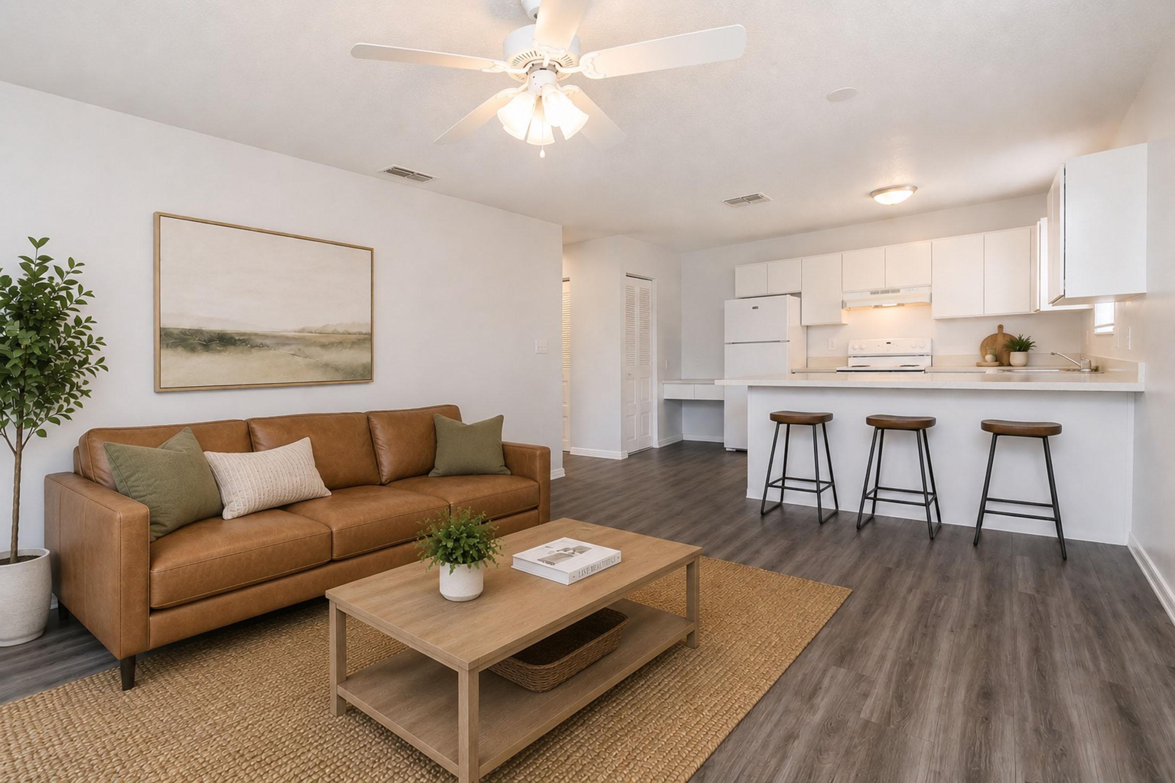 A modern living room featuring a brown leather sofa, a wooden coffee table with a plant, and a neutral rug. In the background, there is a kitchen area with white cabinets and appliances, along with three bar stools at a counter. Soft lighting and light-colored walls create a warm atmosphere.