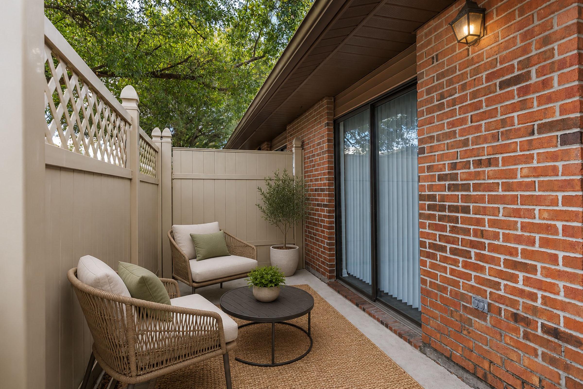 A cozy outdoor sitting area featuring two comfortable chairs, a round coffee table, and a small potted plant, surrounded by a wooden fence and brick walls. Large sliding glass doors lead indoors, and the space is enhanced by natural greenery overhead and a woven rug underfoot.