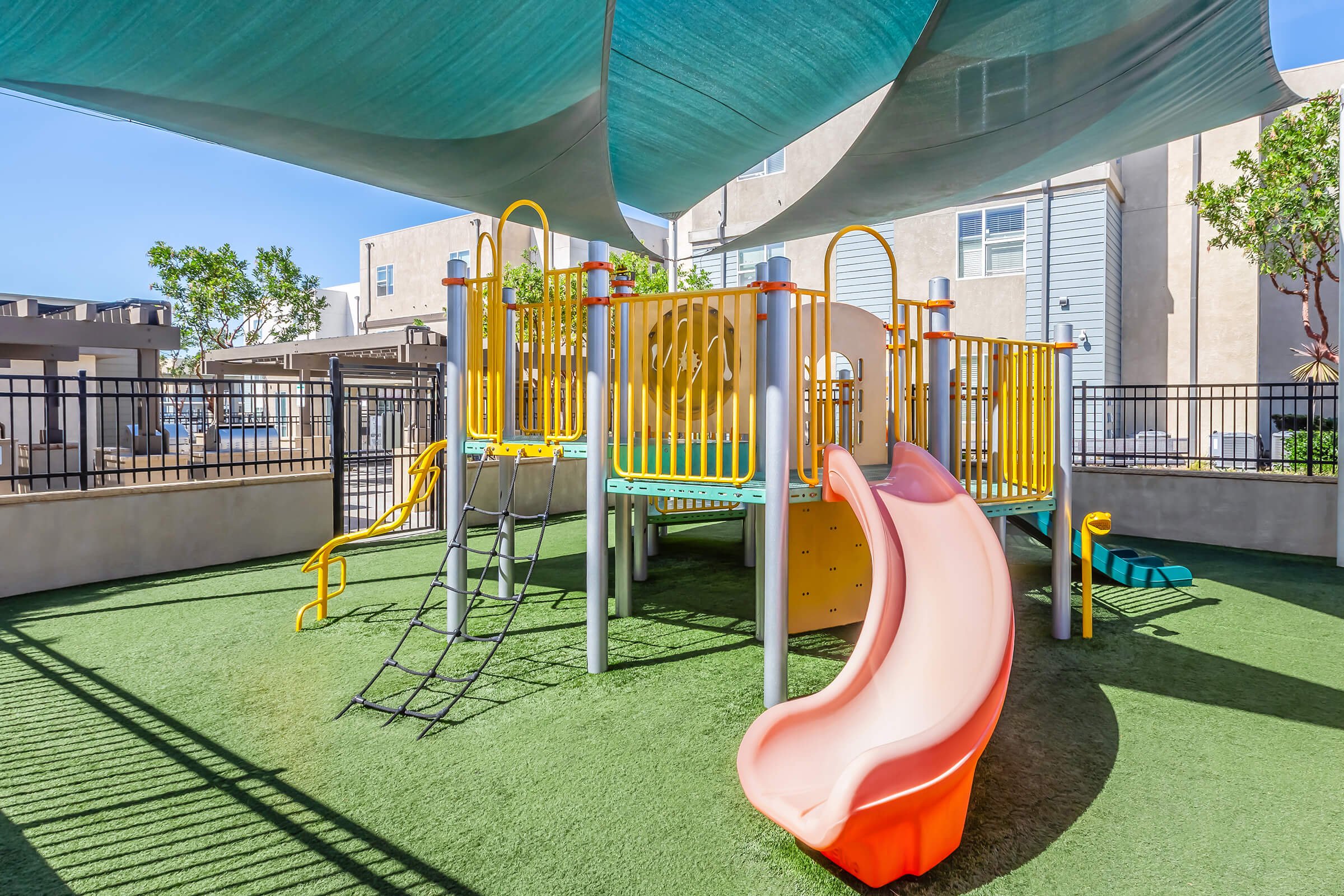 A colorful playground featuring a yellow and orange play structure with slides, climbing frames, and safety railings, situated under a shaded canopy. The ground is covered in green grass-like material, creating a safe space for children to play. In the background, there are some buildings and trees.
