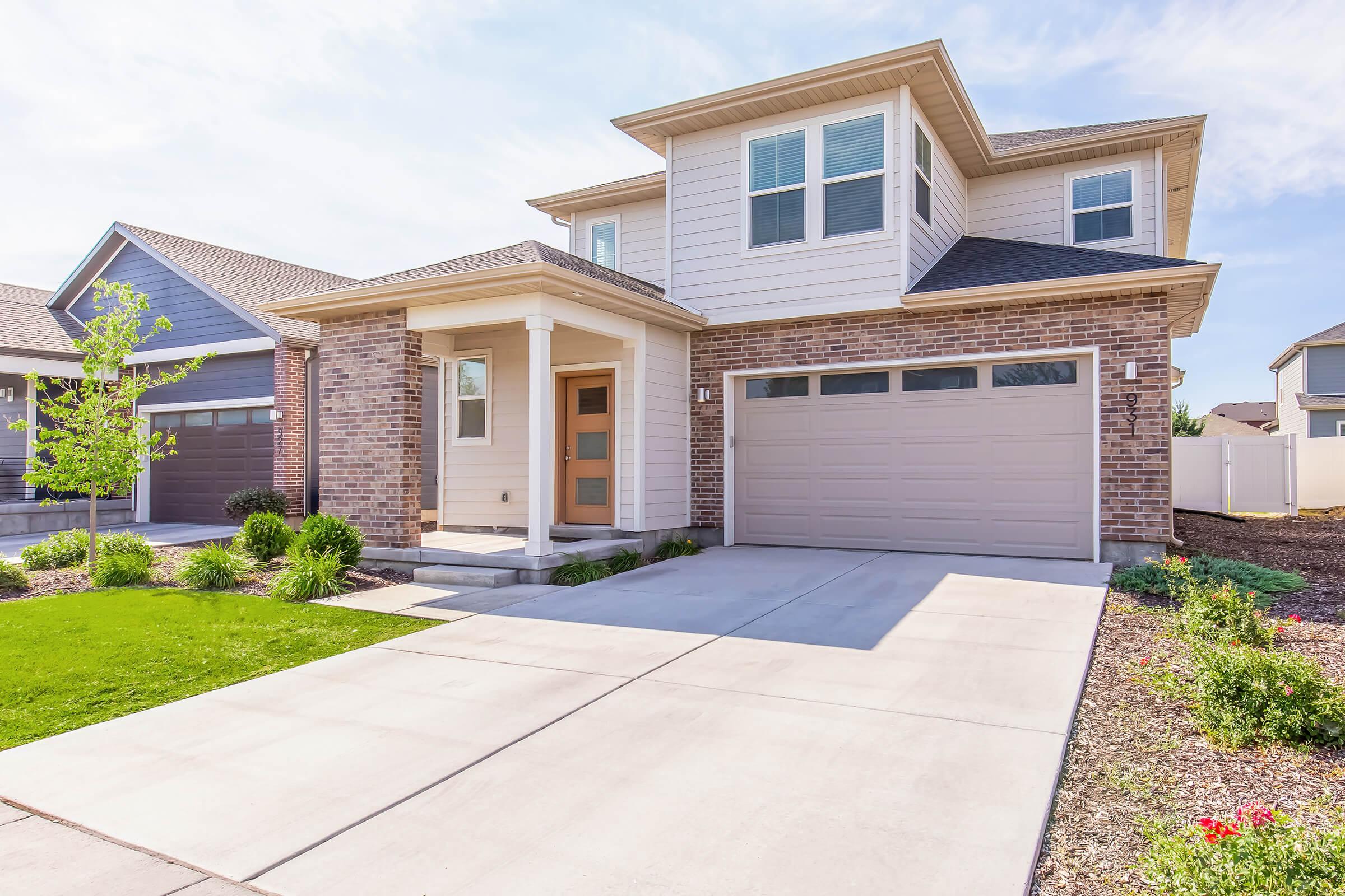 A modern two-story house with a brick and siding exterior. It features a welcoming front porch, large windows, and a two-car garage. The driveway is paved, with neatly landscaped grass and shrubbery in the front yard. Bright blue sky and soft clouds are visible in the background.