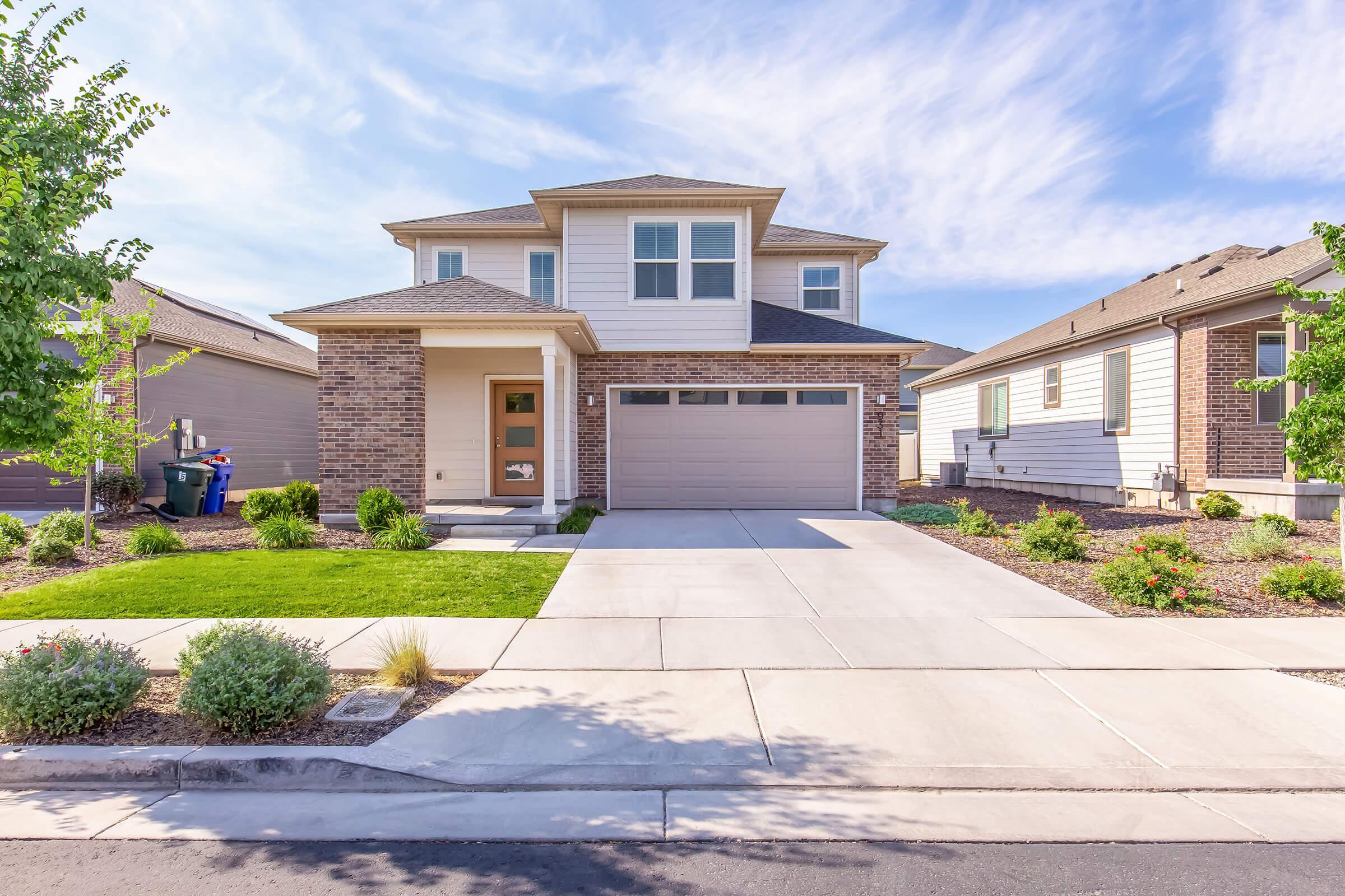 A modern two-story house with a brick front, a two-car garage, and landscaped front yard, including shrubs and small trees. The driveway is paved, and the house features large windows and a welcoming porch. Nearby, there are additional homes visible on either side.