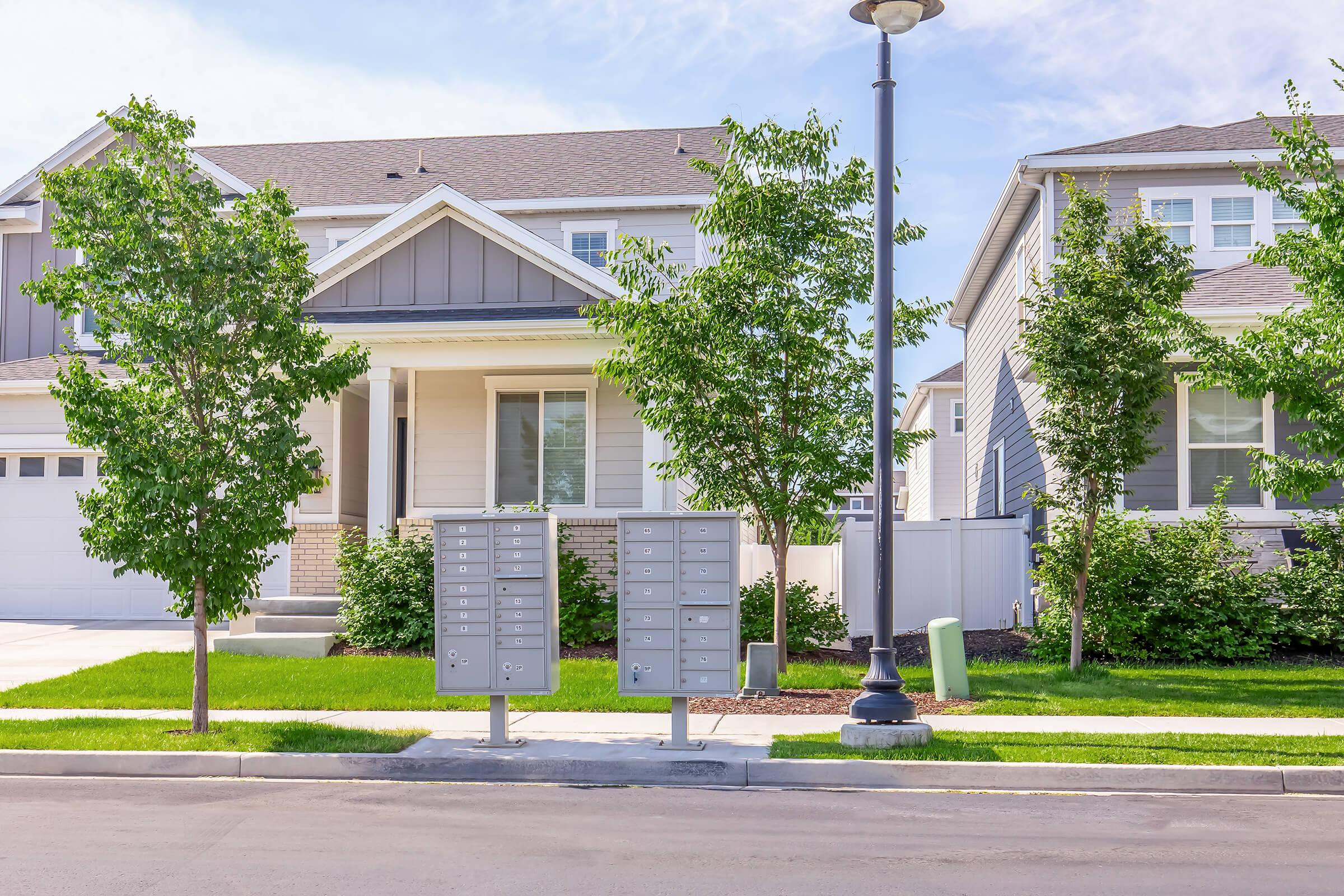 A residential area featuring two identical multi-unit mailboxes on a sidewalk, surrounded by manicured lawns and trees. In the background, there are two modern homes with gray siding, a garage, and a light post. The scene is bright and sunny, with blue skies and a well-maintained neighborhood appearance.