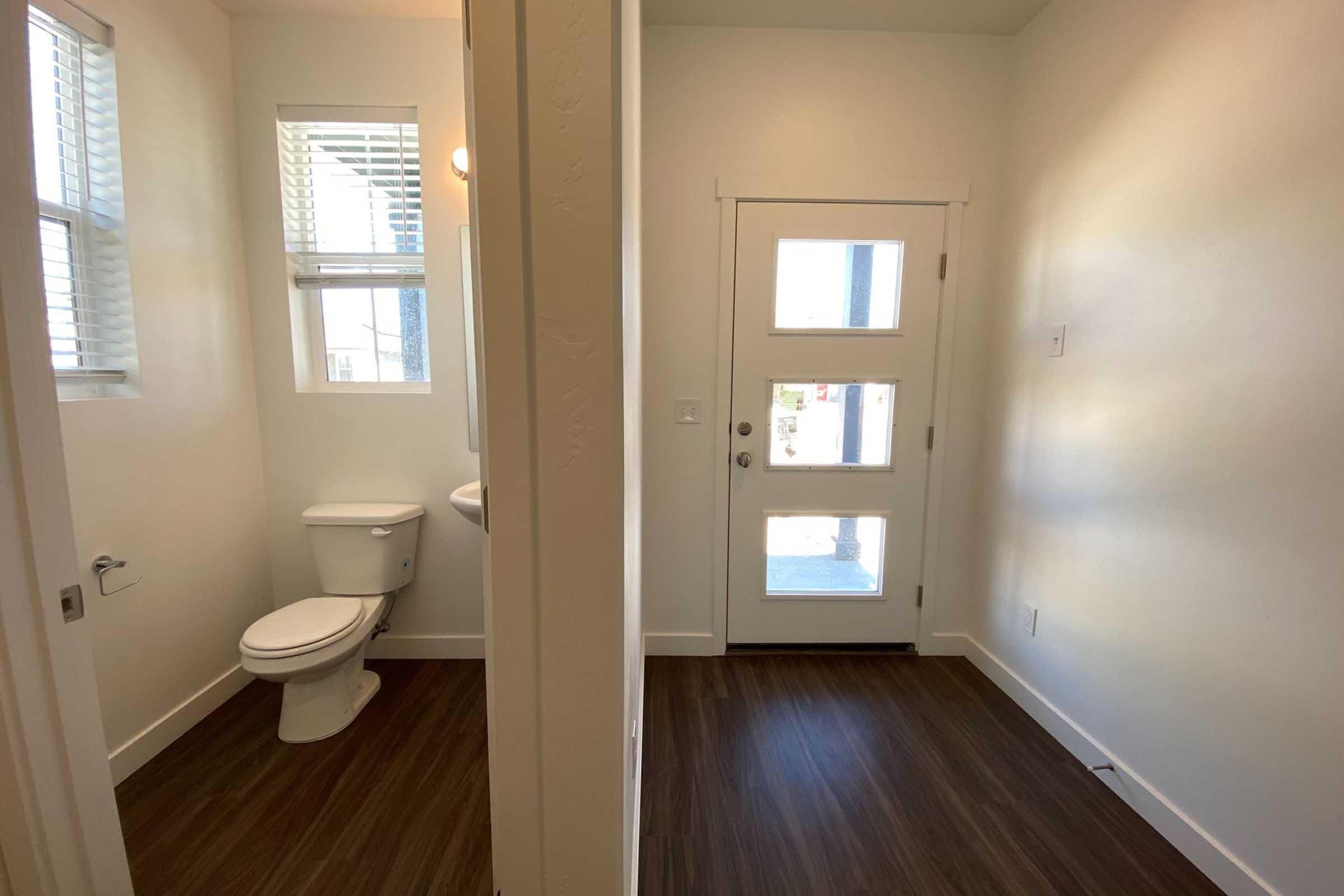 A well-lit hallway leading to a door, with a small bathroom area on the left featuring a toilet and a window. The flooring is dark wood, and the walls are painted white, creating a modern and clean aesthetic. The door at the end of the hallway has large glass panels.