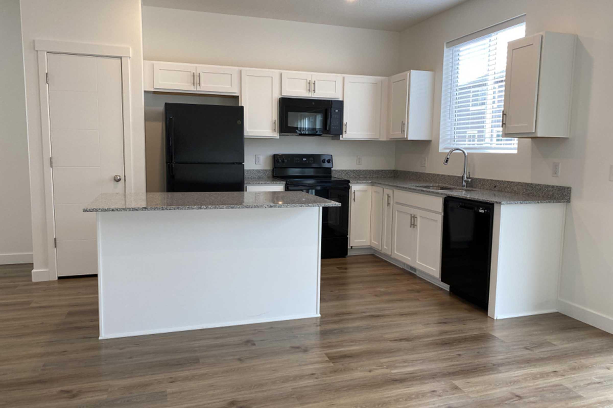 A modern kitchen featuring white cabinets, a black refrigerator, a microwave, and a stove. The countertops are gray with a speckled pattern. A large window allows natural light in, illuminating the wood-style flooring. The layout includes a kitchen island with additional seating space.