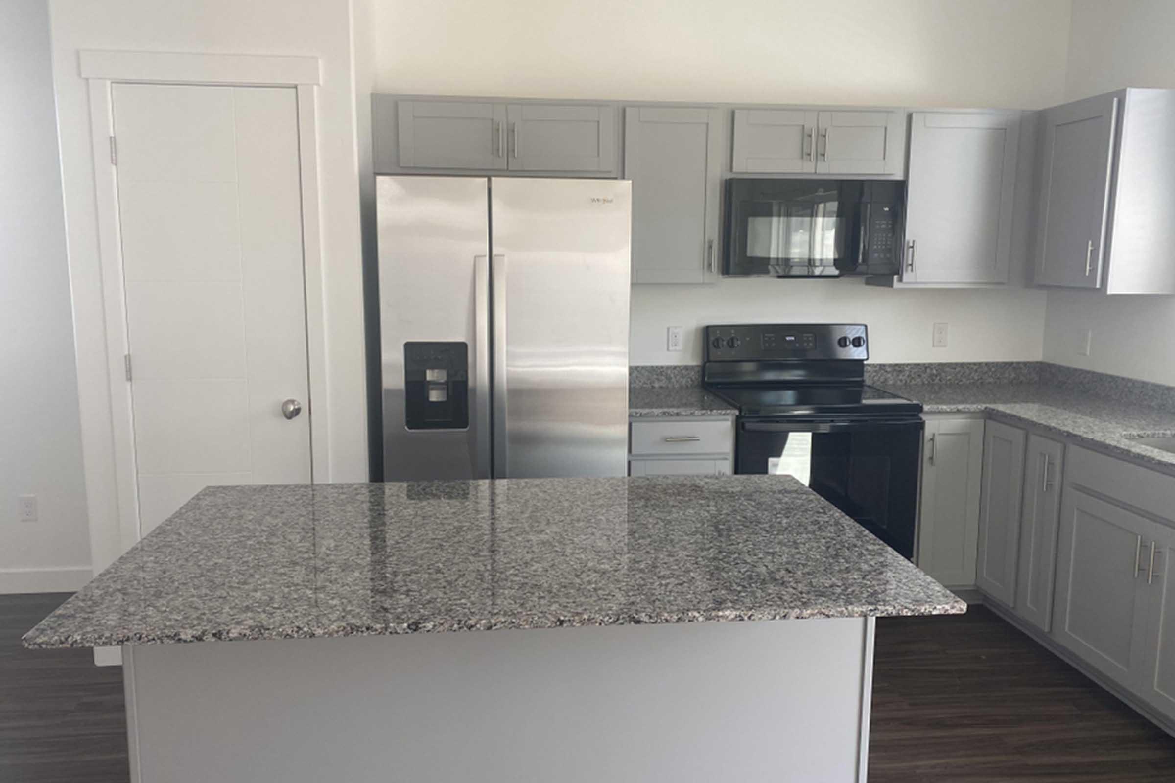 A modern kitchen featuring stainless steel appliances, including a refrigerator, microwave, and oven. The cabinetry is light gray, and there's a large granite countertop island in the center. The floor has a dark wood finish, and the walls are painted in a light color, creating a bright and inviting space.
