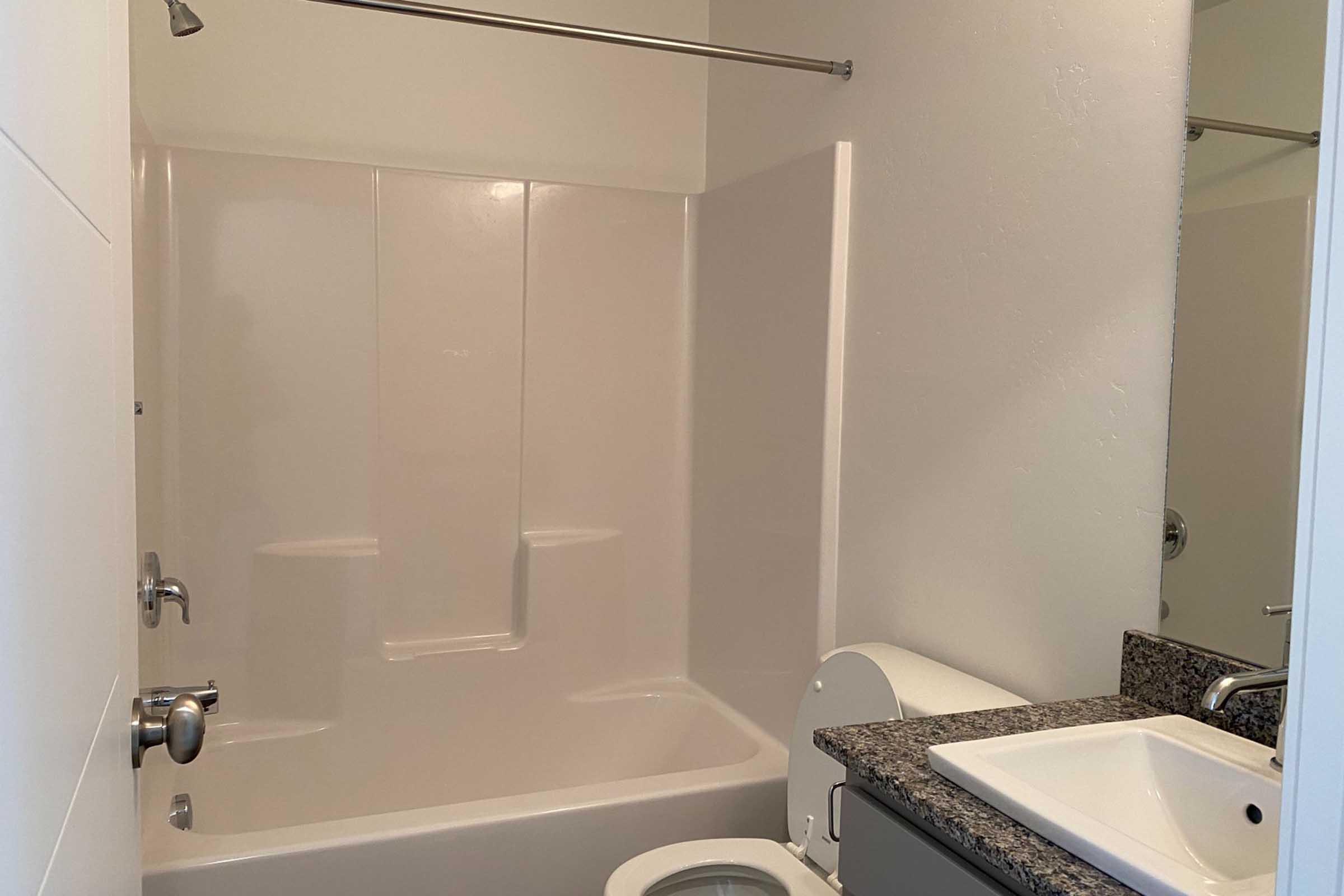 A clean, modern bathroom featuring a white bathtub with a showerhead, a toilet, and a sink with a dark granite countertop. The walls are painted in a light color, enhancing the bright and spacious feel of the room. The layout is functional and minimalist, with chrome fixtures.