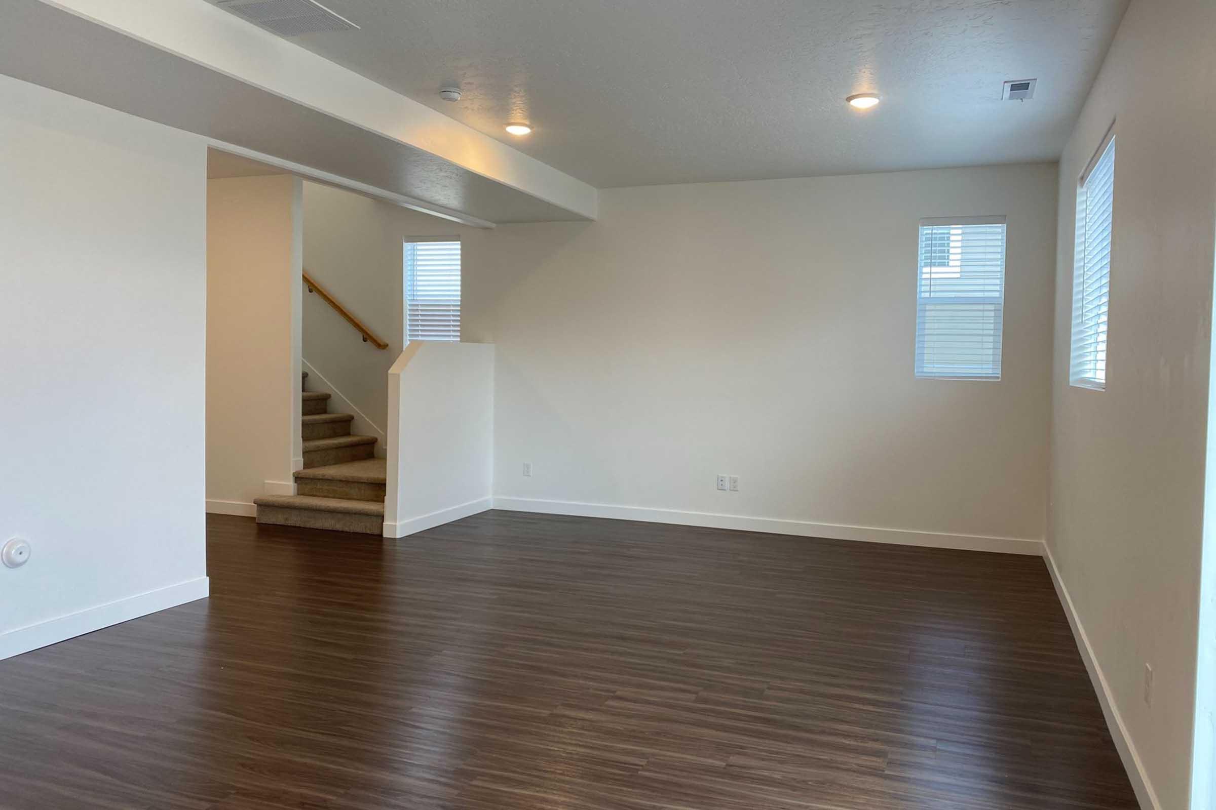 A spacious, empty room with modern decor featuring hardwood flooring, white walls, and natural light from two small windows. A staircase is visible in the background, leading to the upper level. The room is clean and unfurnished, creating a blank canvas for potential decoration or use.
