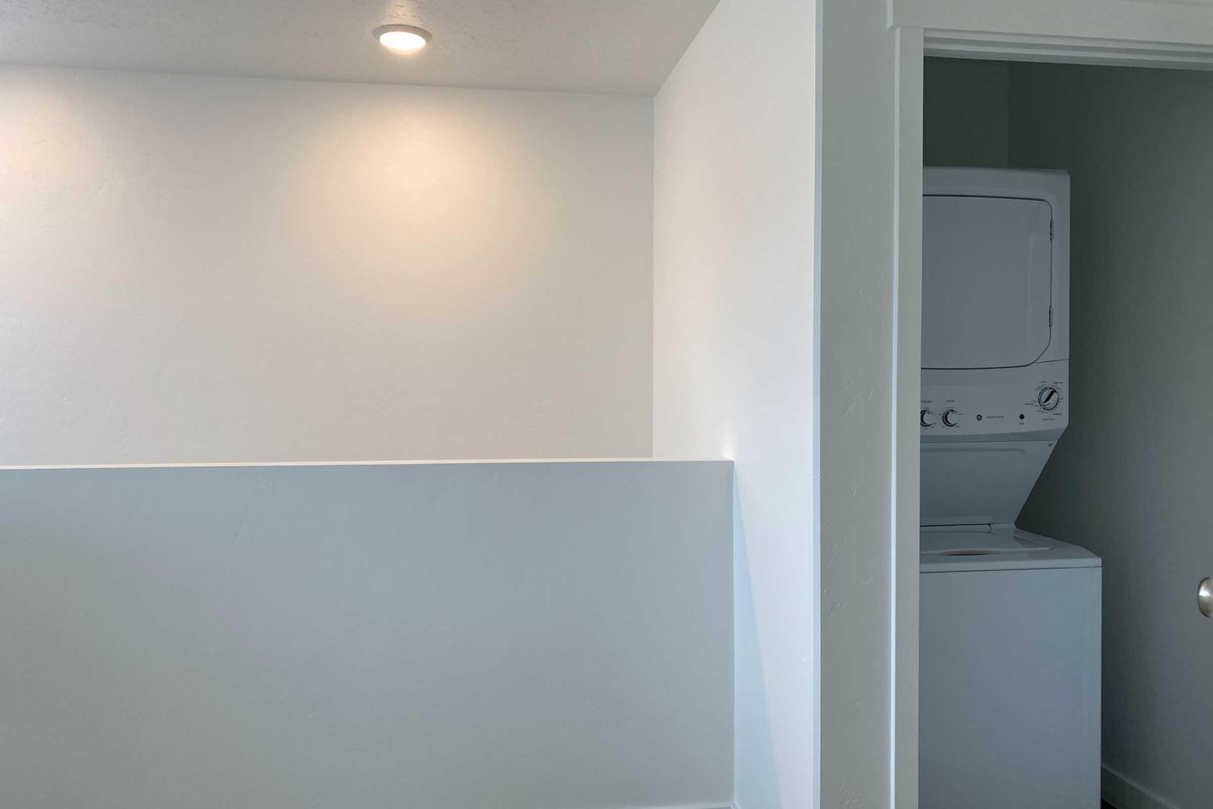 Interior of a laundry room featuring a white wall with recessed lighting and a stacked washing machine and dryer. A doorway is visible on the right, leading to another room. The space is bright and minimalistic, emphasizing cleanliness and functionality.